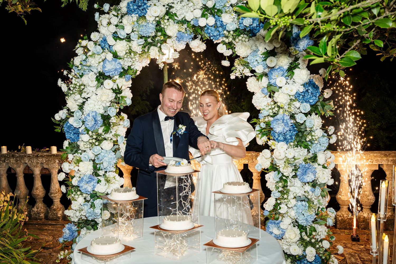 Elegant wedding cake cutting ceremony under floral arch with blue and white hydrangeas at night.