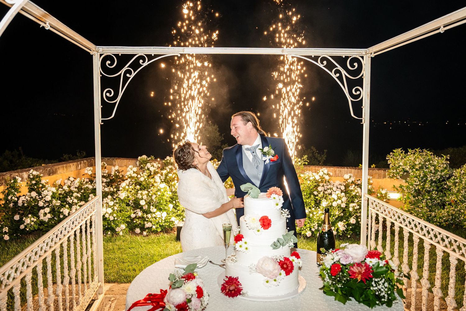 Bride and groom kiss while cutting wedding cake under gazebo with spectacular fireworks display.