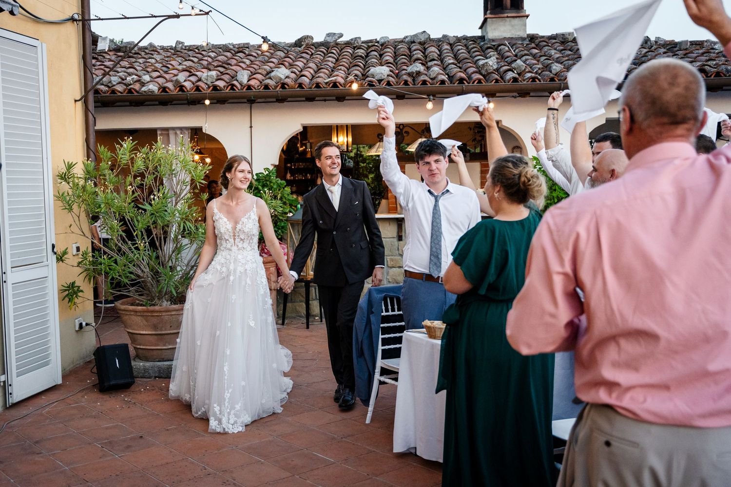 Bride and groom enter outdoor wedding reception as guests celebrate with raised hands and white napkins.