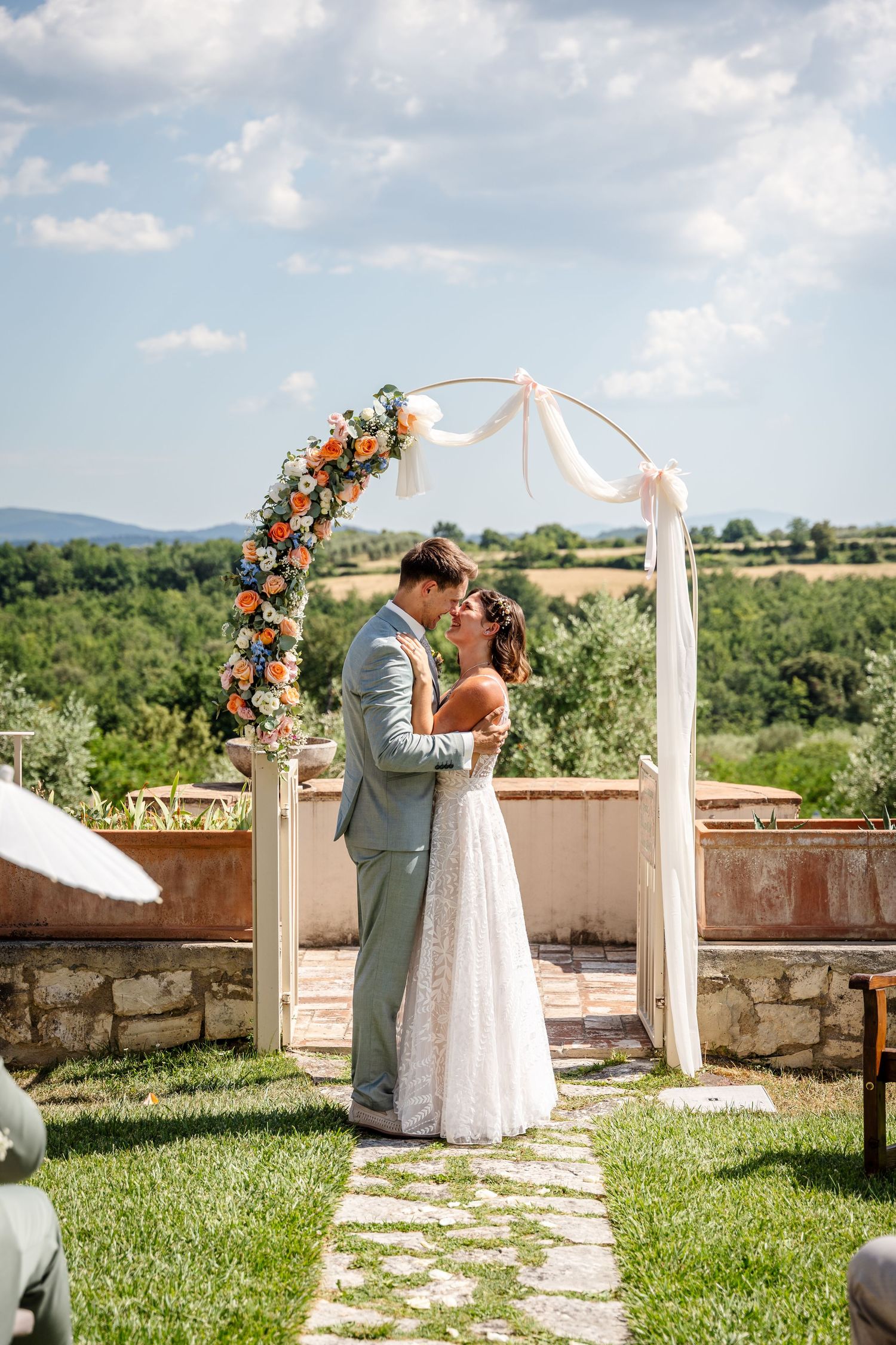 Bride and groom share a romantic moment under a floral arch with orange and blue flowers at an outdoor Tuscan wedding.