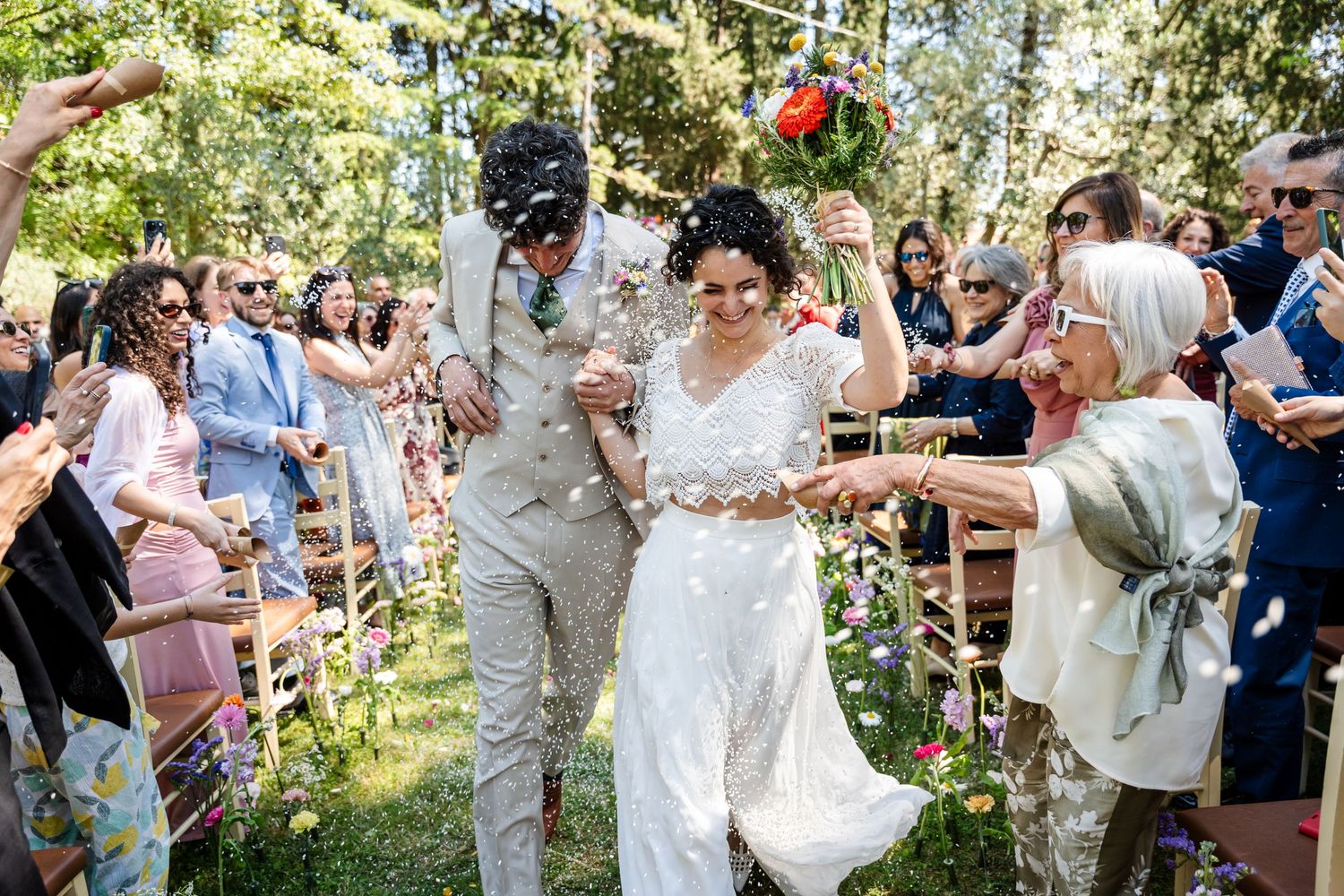 Joyful wedding celebration outdoors with guests throwing flower petals as couple walks down garden path.