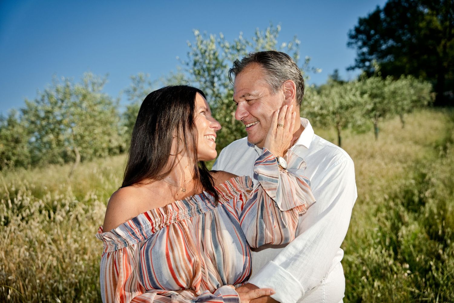 A smiling couple embraces outdoors in a sunny green field, gazing at each other affectionately on a clear summer day.