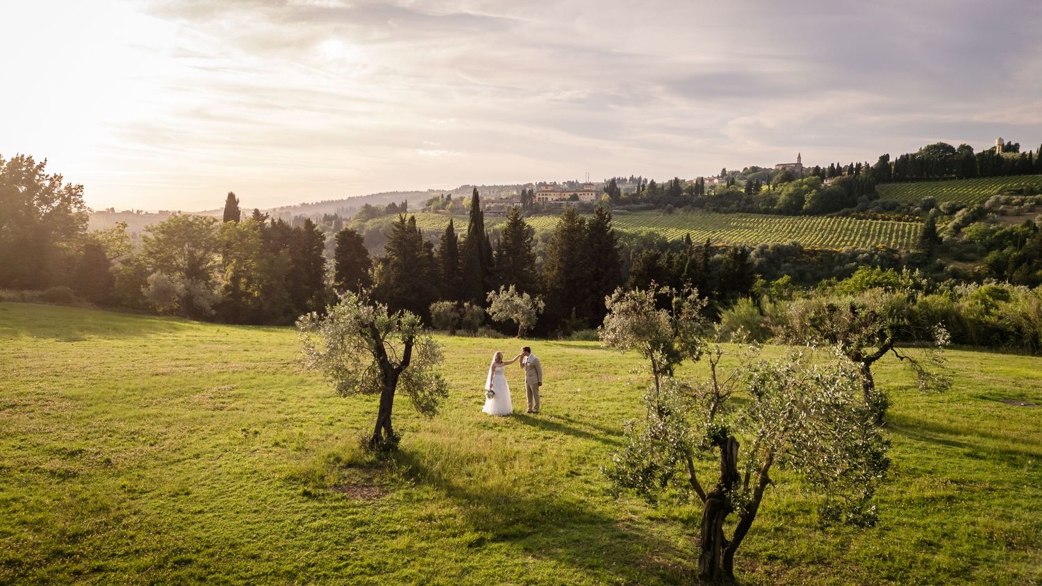 Aerial view of a wedding couple walking through a lush green Tuscan countryside with cypress trees at golden sunset.