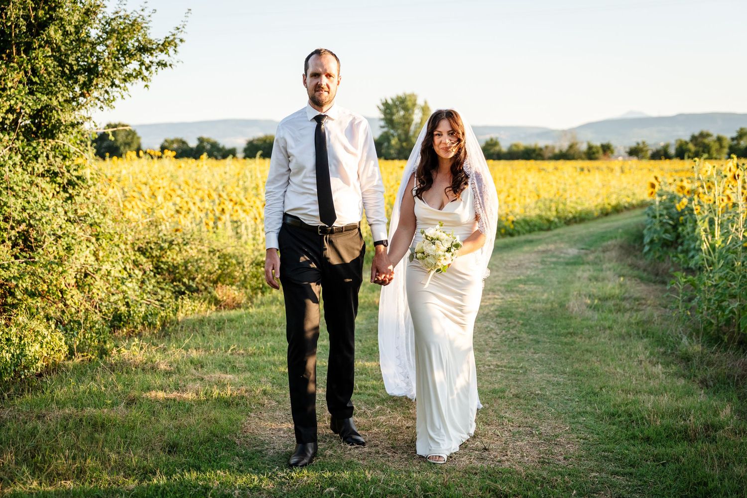 Bride and groom holding hands walking along a sunflower field path on their wedding day.