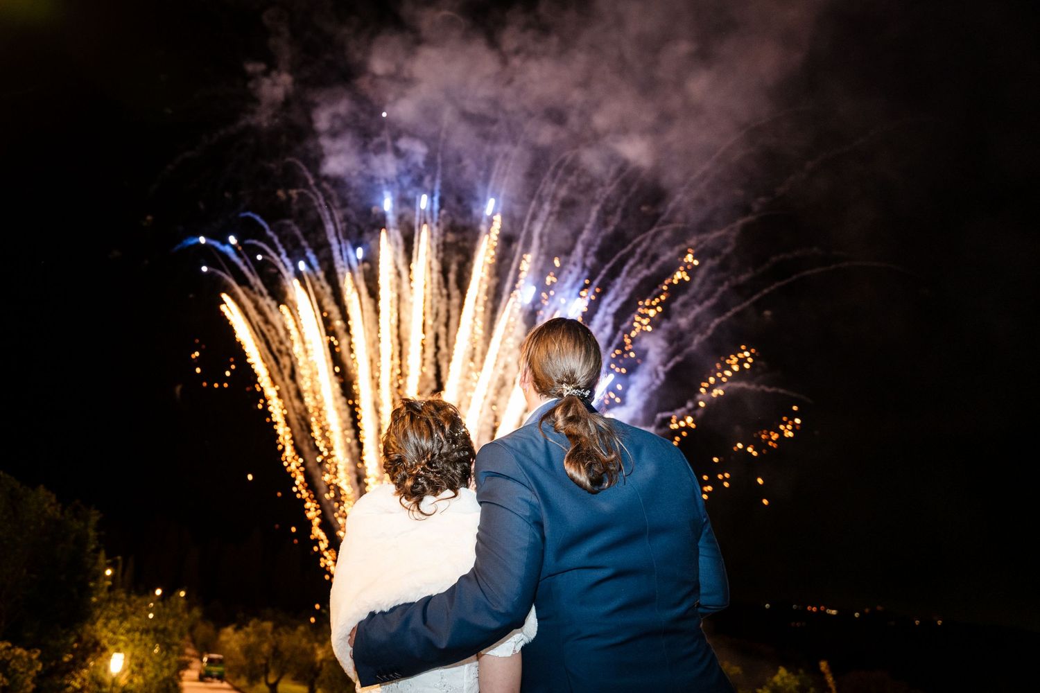 A couple watches a spectacular fireworks display at night while embracing in their wedding attire.