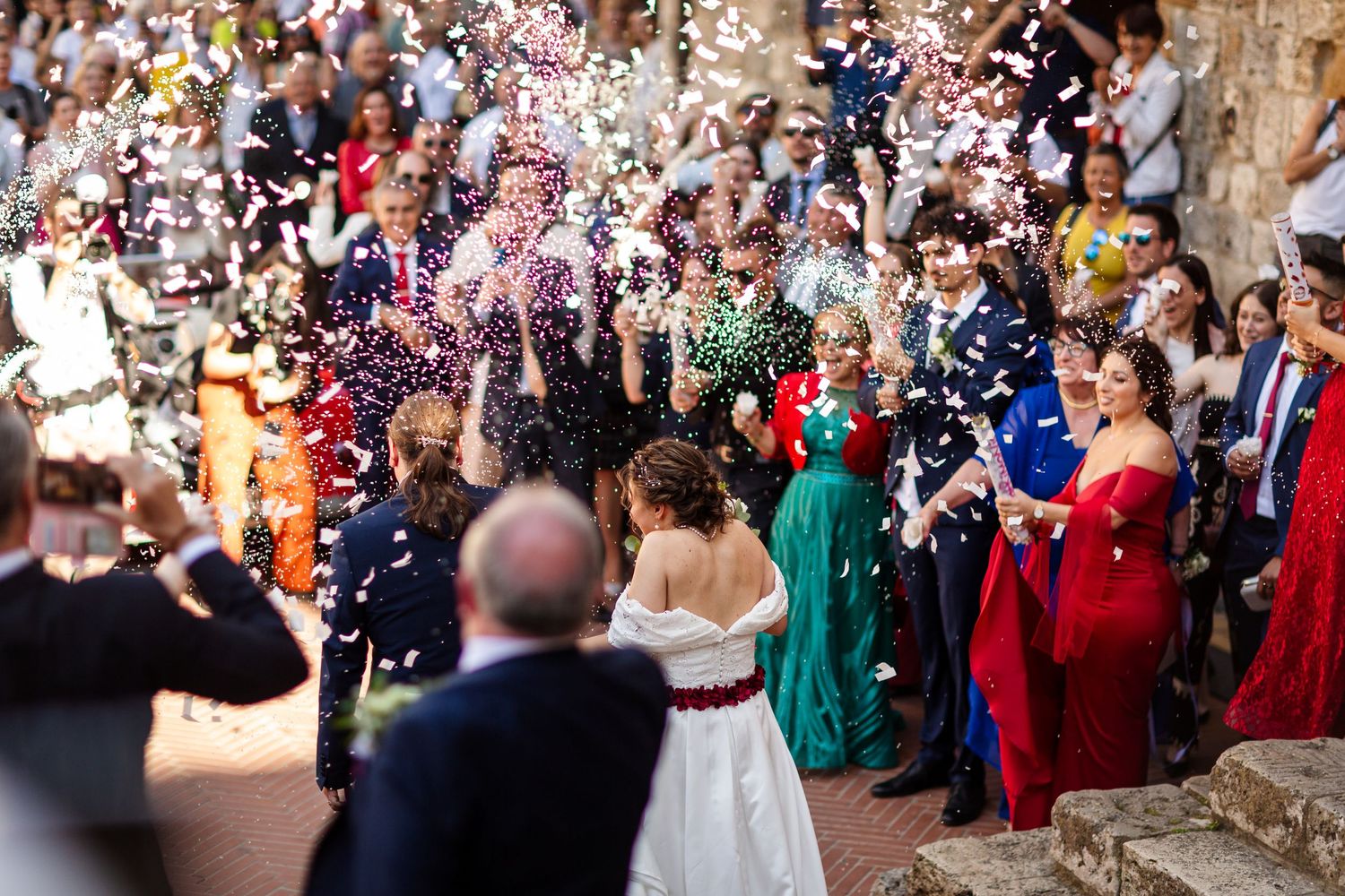 Joyful wedding guests throwing colorful confetti over bride and groom during outdoor ceremony celebration.