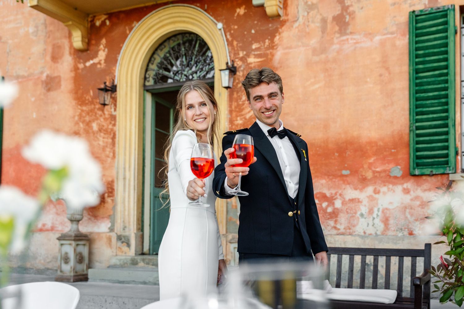 Elegant couple in wedding attire toasting with red wine glasses outside a rustic Italian villa.