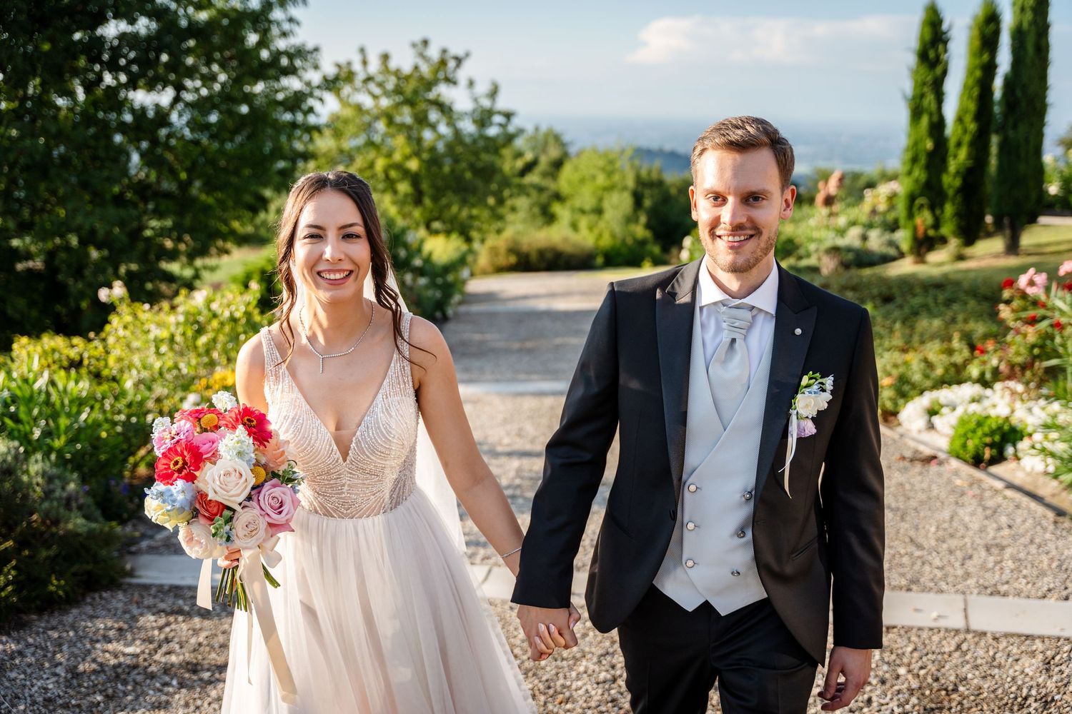 Happy bride and groom walking hand in hand outdoors on their wedding day, bride holding colorful bouquet.