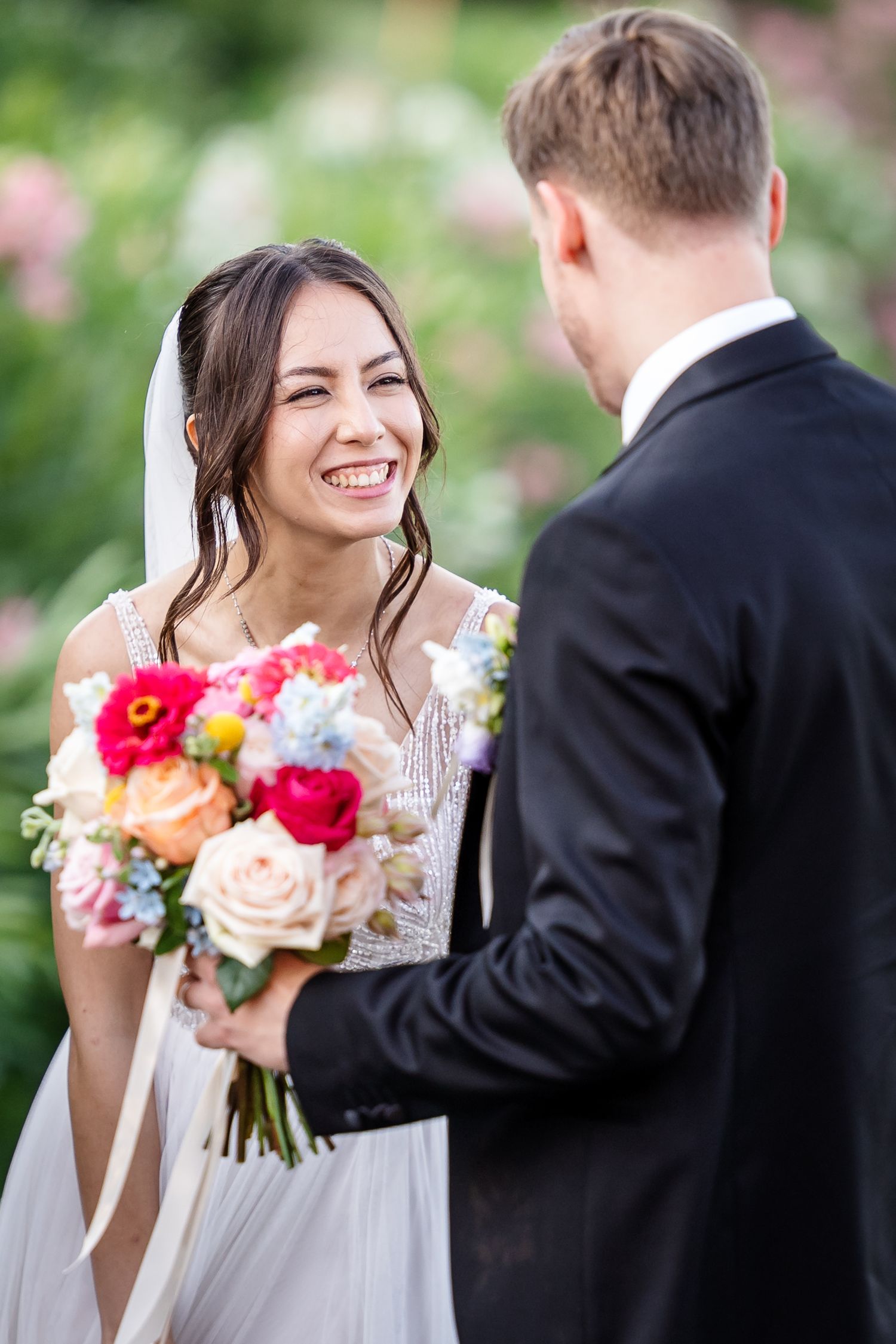 Radiant bride laughing while holding vibrant bouquet facing groom in black suit outdoors.
