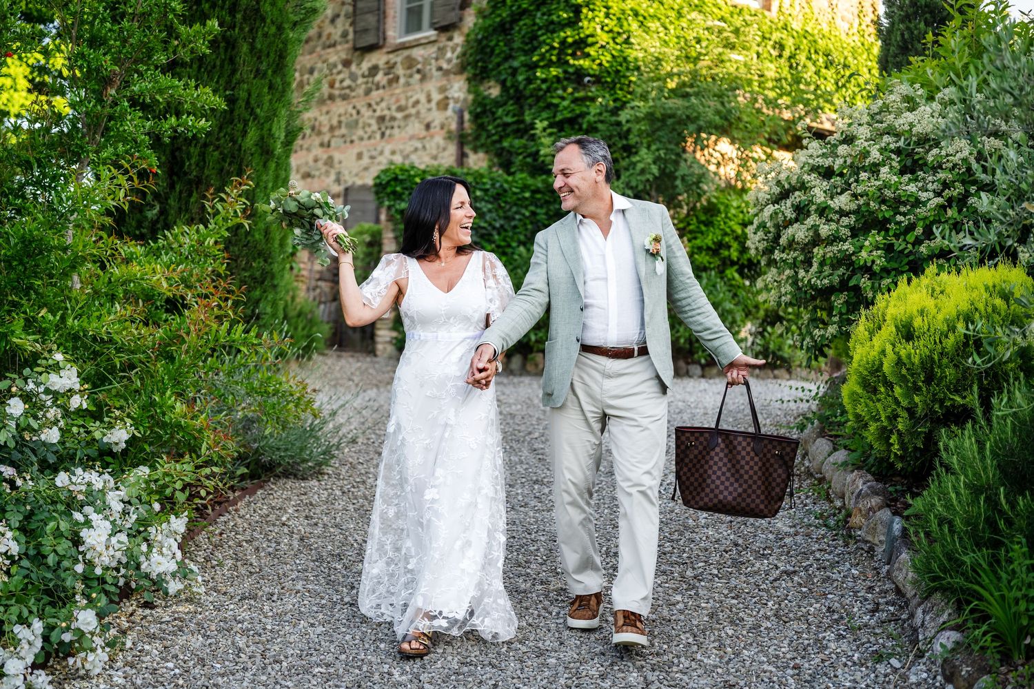 Bride and groom walk hand in hand through a lush garden path surrounded by vibrant green foliage.