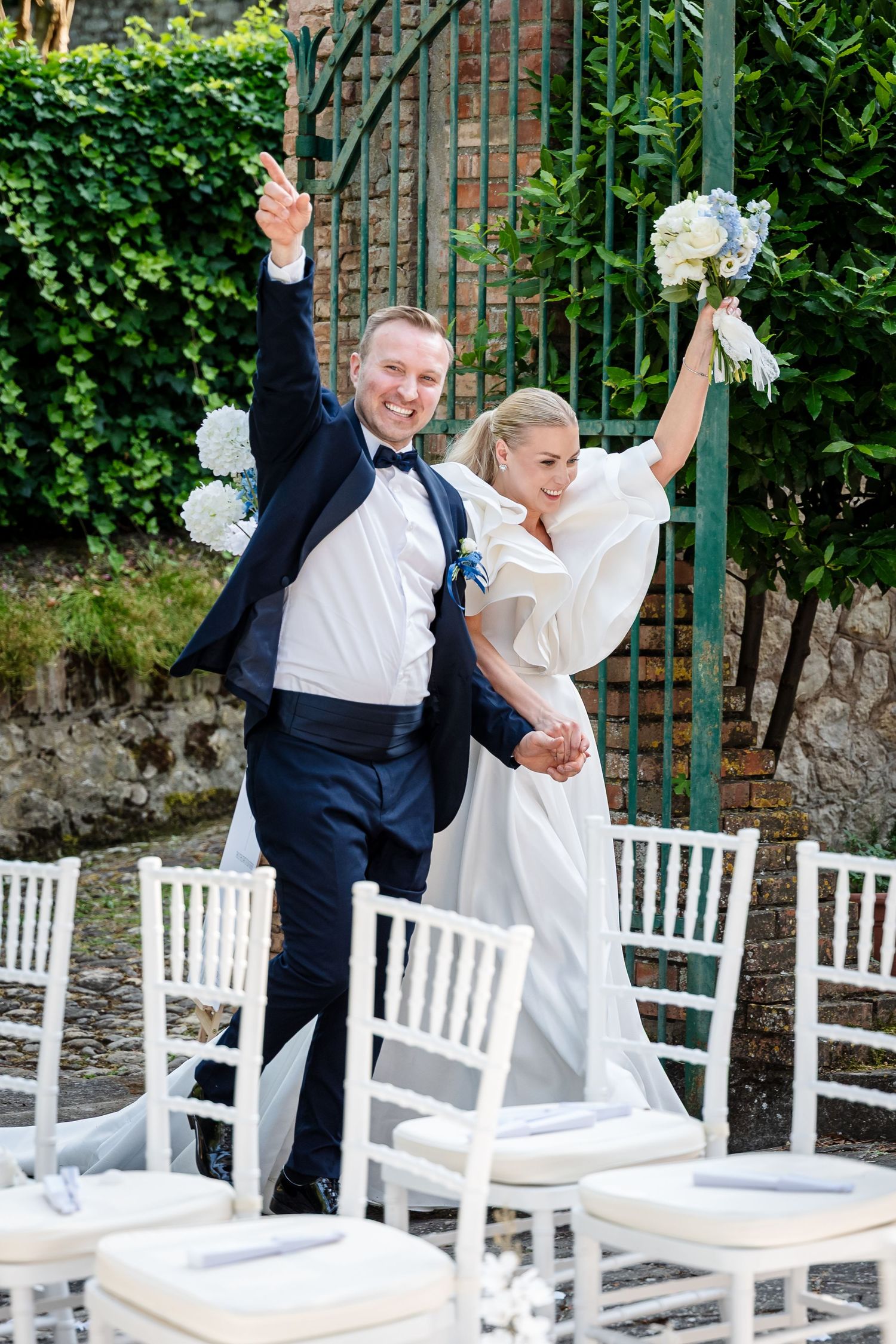 A couple celebrates joyfully in front of white chairs at an outdoor wedding venue with lush greenery in background.