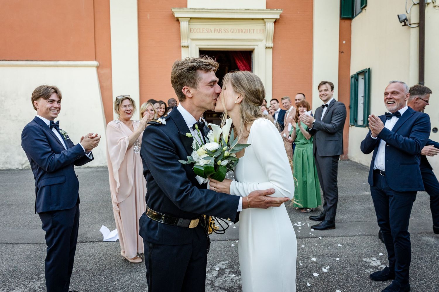 Newlywed couple sharing a kiss outside a church while wedding guests clap and celebrate around them.