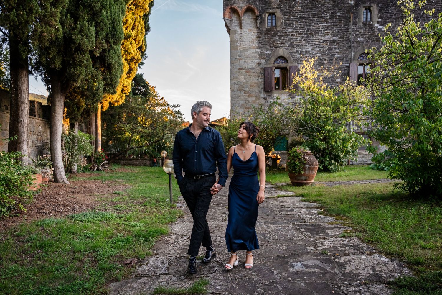 Couple walking hand in hand through a lush Italian castle garden at sunset in elegant evening attire.