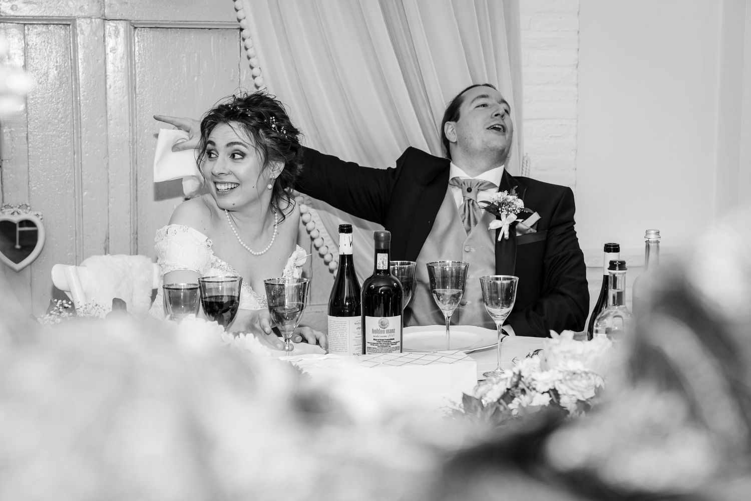Black and white candid photo of laughing bride and groom seated at wedding reception table.