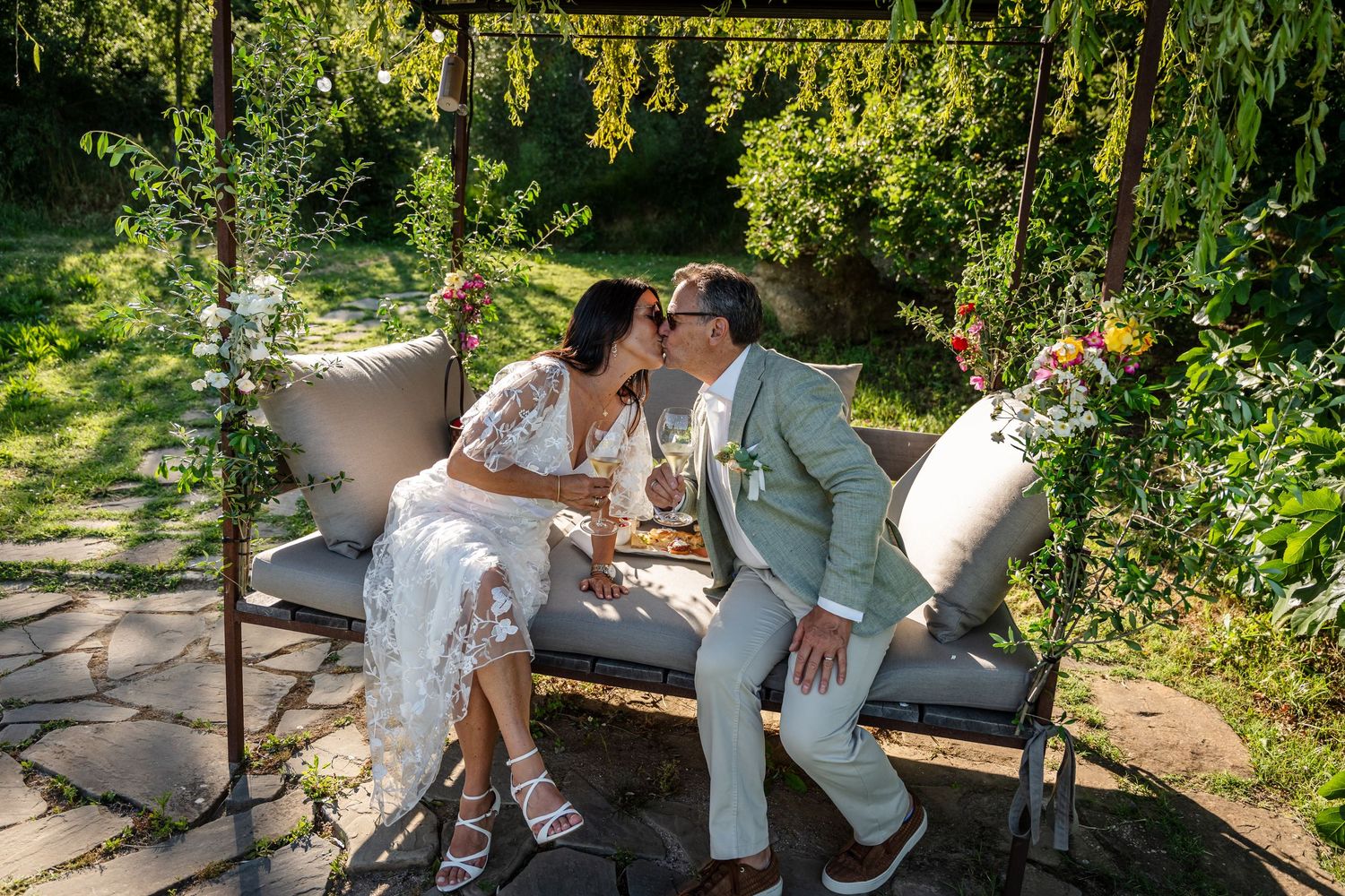 Couple in wedding attire share an intimate moment on a garden bench surrounded by lush greenery on a sunny day.