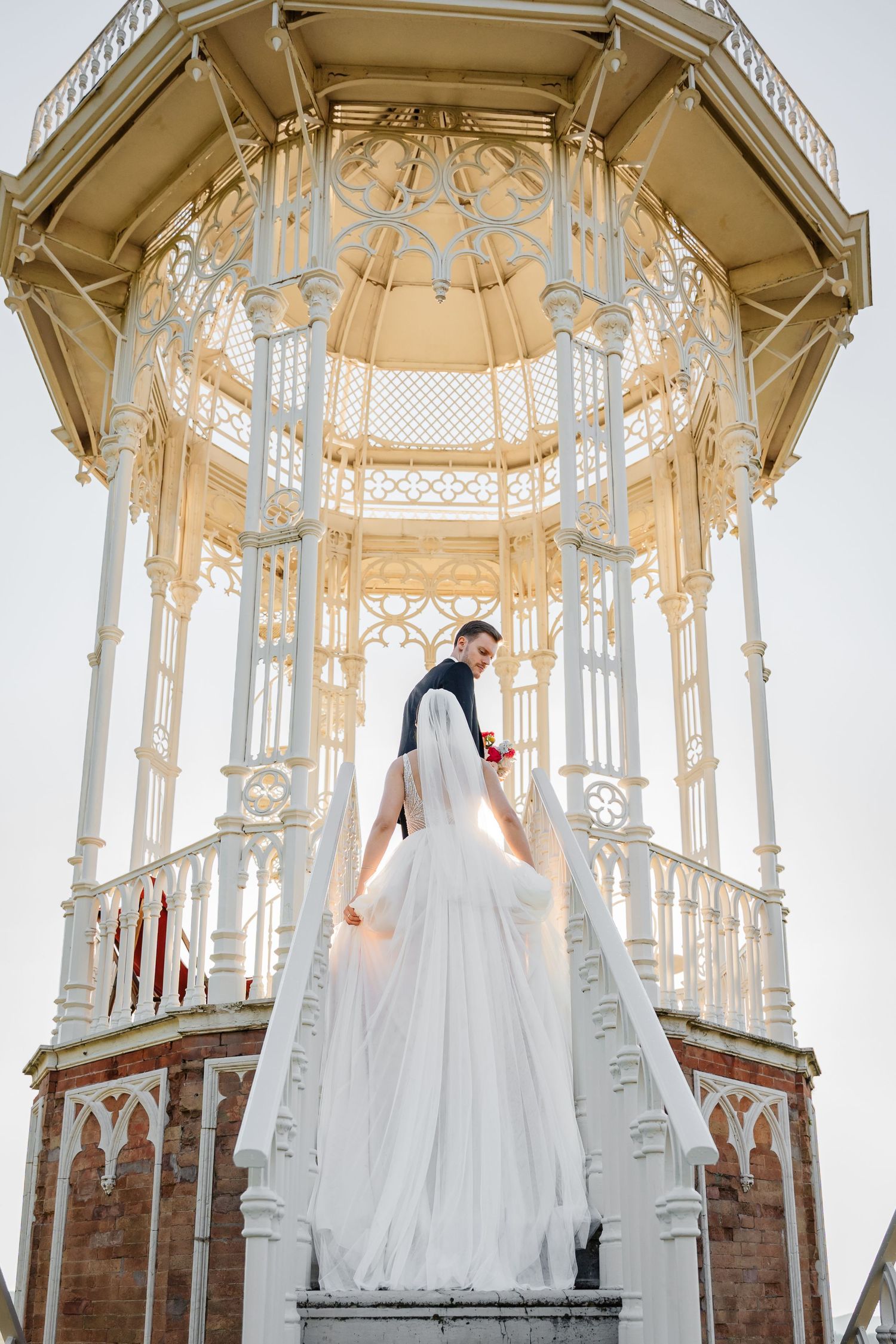 Bride and groom stand at ornate white gazebo entrance, bride in flowing white gown with long veil, romantic wedding photo.