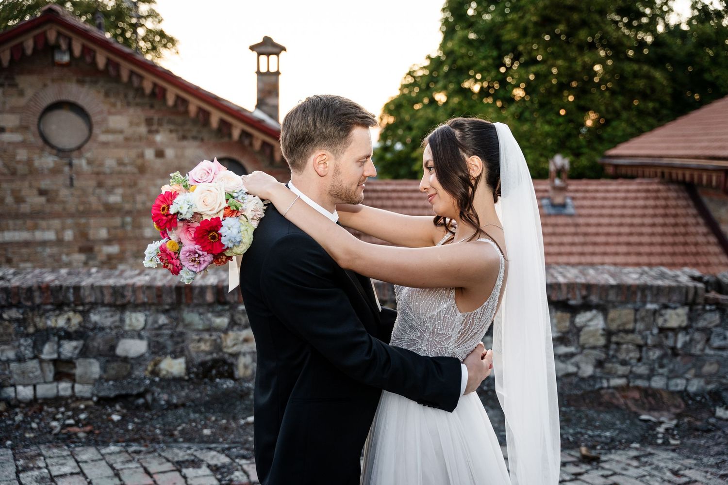 Romantic bride and groom embrace holding a bouquet near a rustic stone wall at dusk.