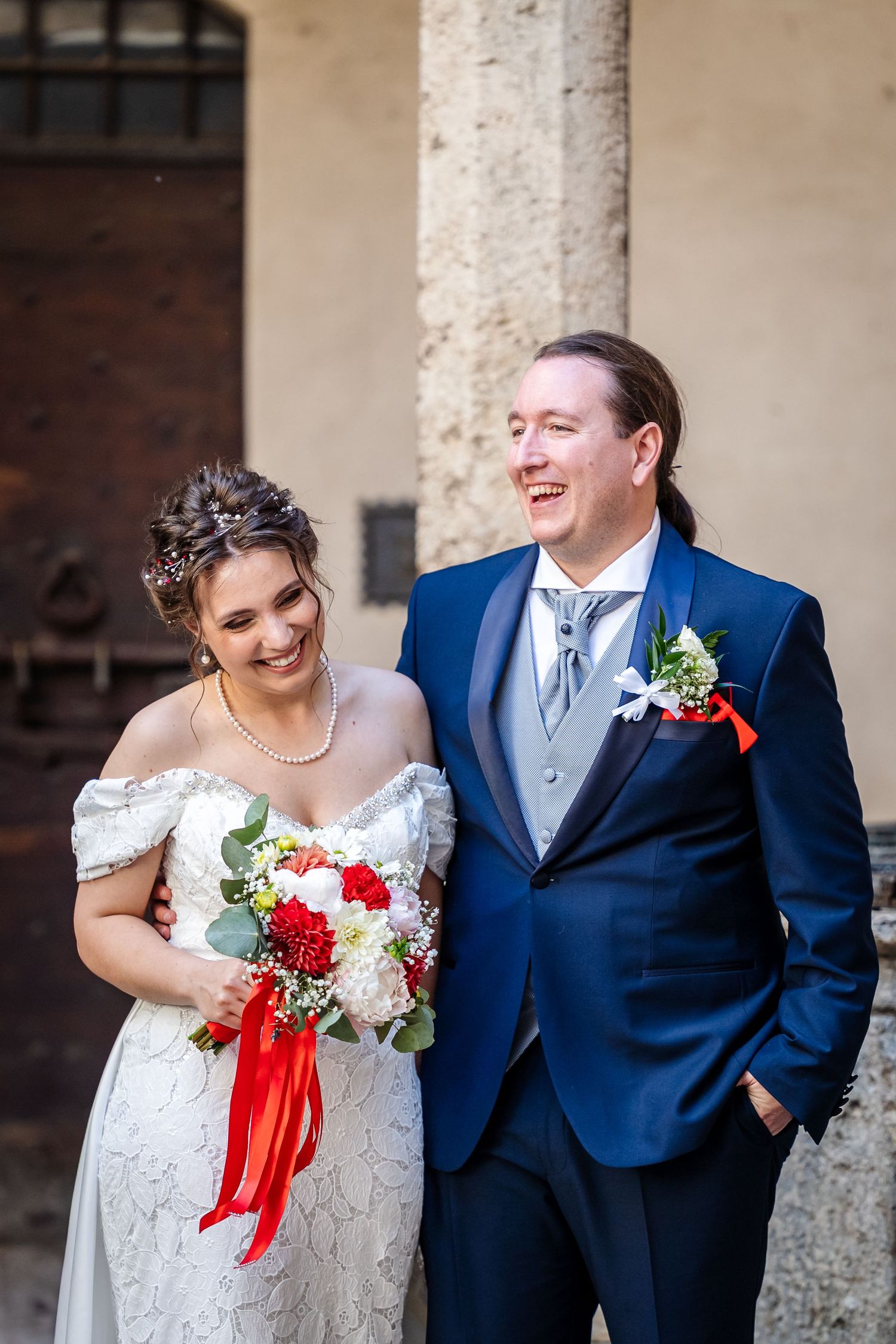 Smiling bride and groom in blue suit pose together outside historic building on their wedding day.