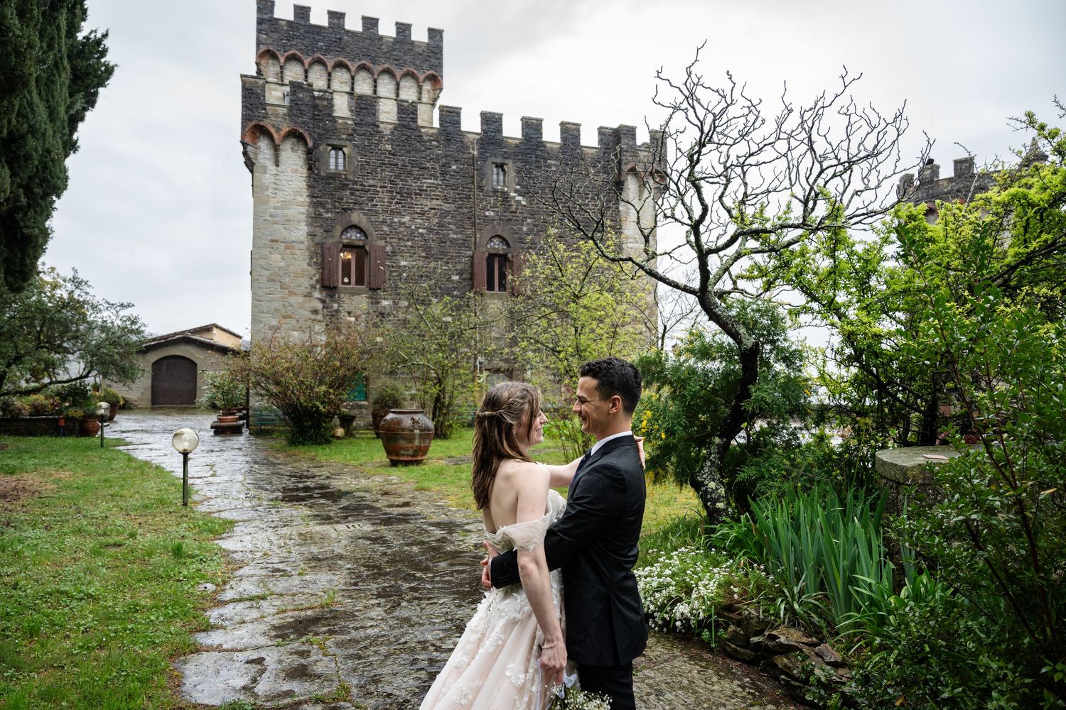 A couple stands on a wet stone path outside a medieval castle tower in the rain during a romantic celebration.