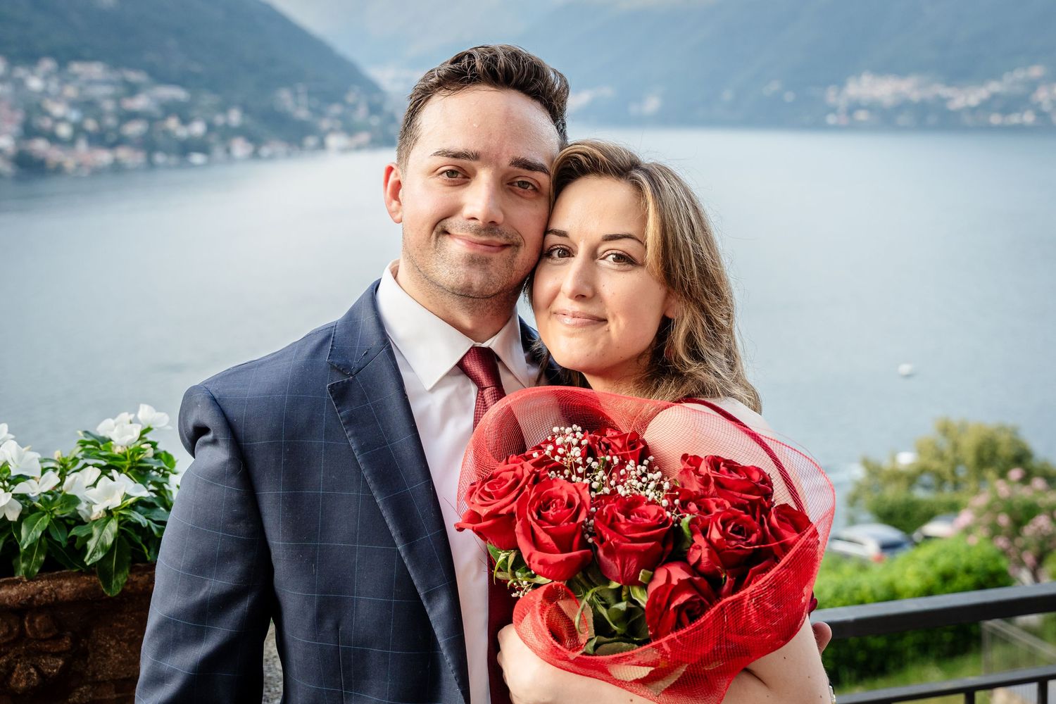 A couple poses for wedding photos against a scenic mountain lake backdrop with red rose bouquet and formal attire.