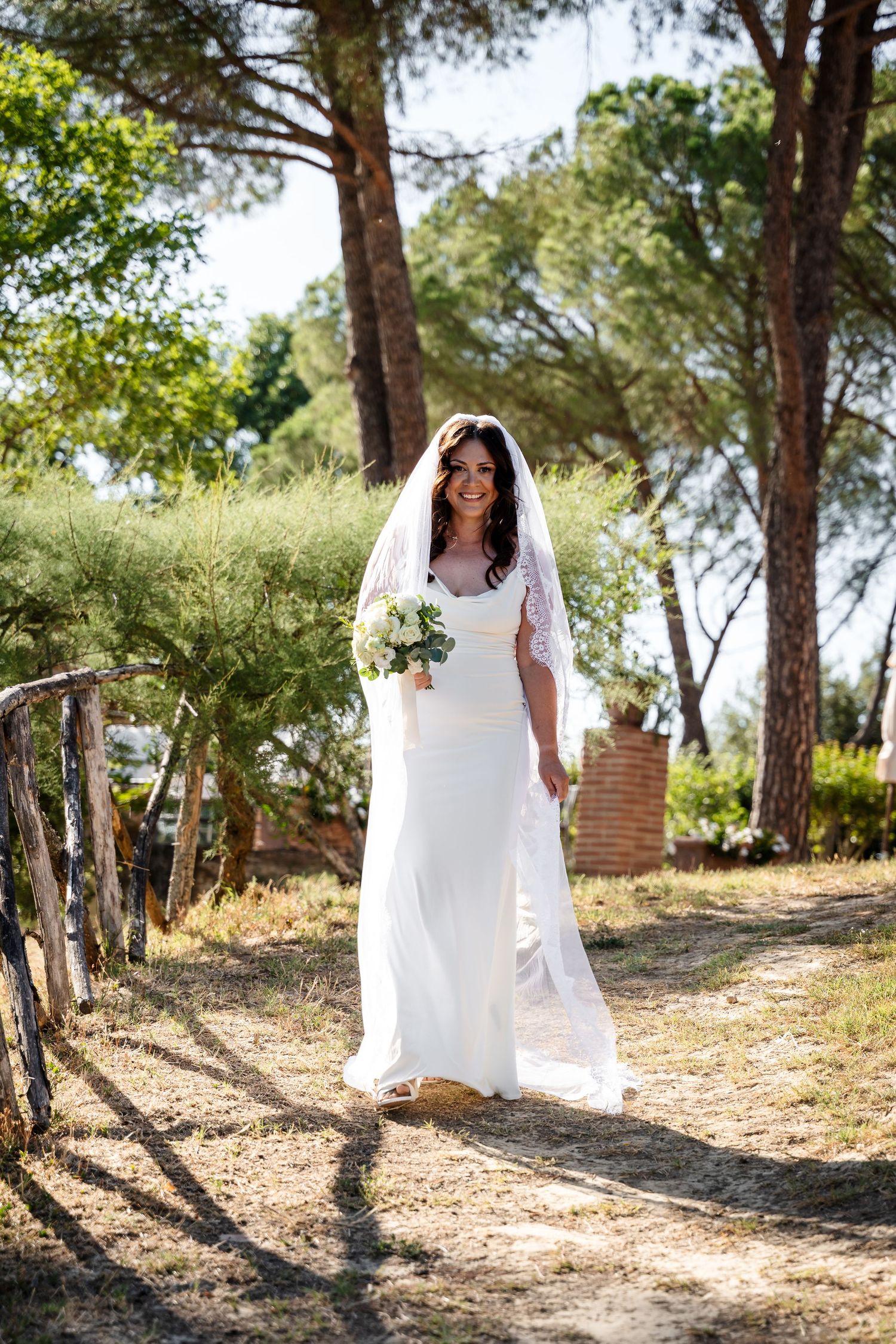 A bride in a flowing white dress walks through a sunlit forest setting, creating a dreamy outdoor bridal portrait series.