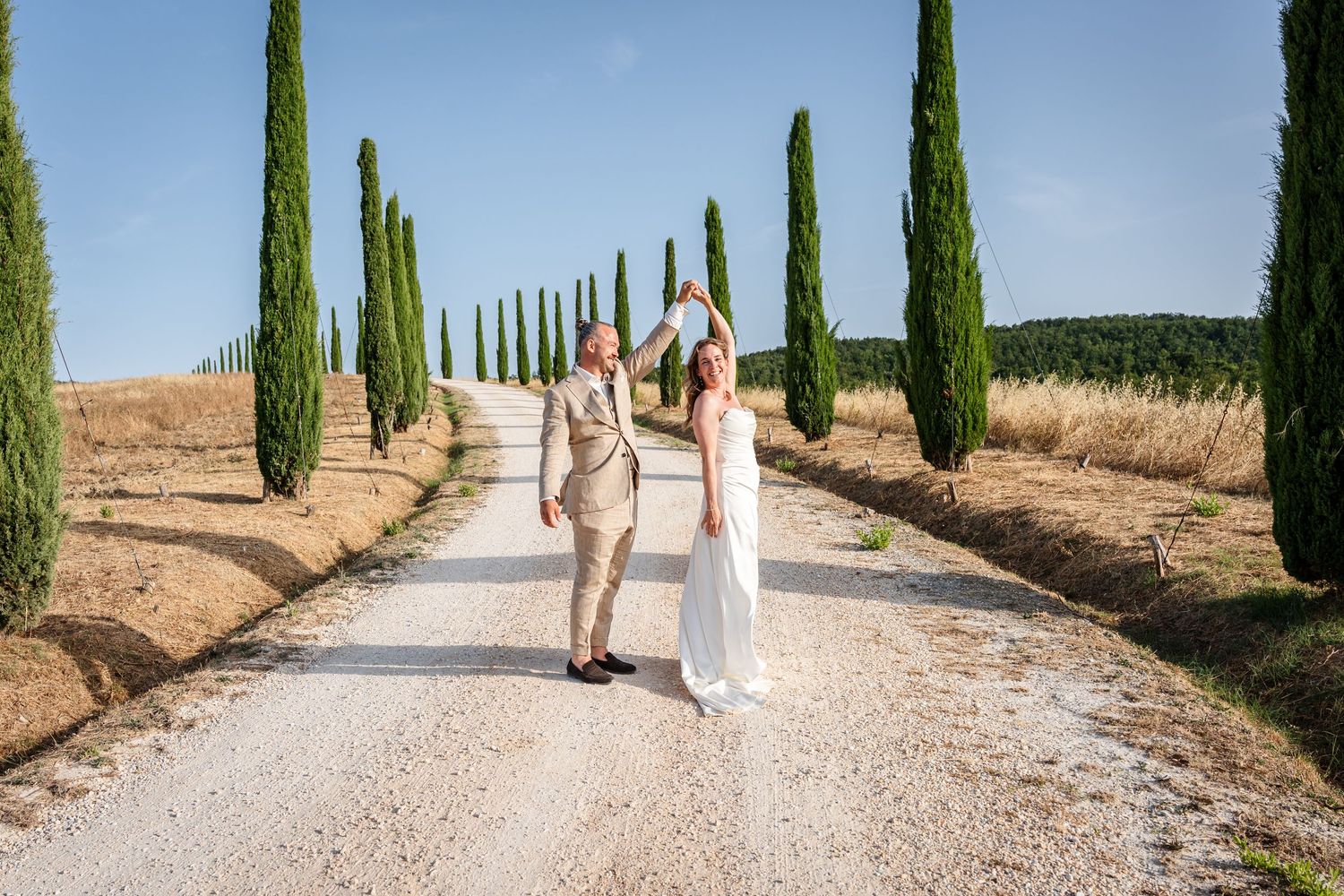 Couple in wedding attire walking down a cypress-lined gravel path in Tuscany, Italy on a sunny day.