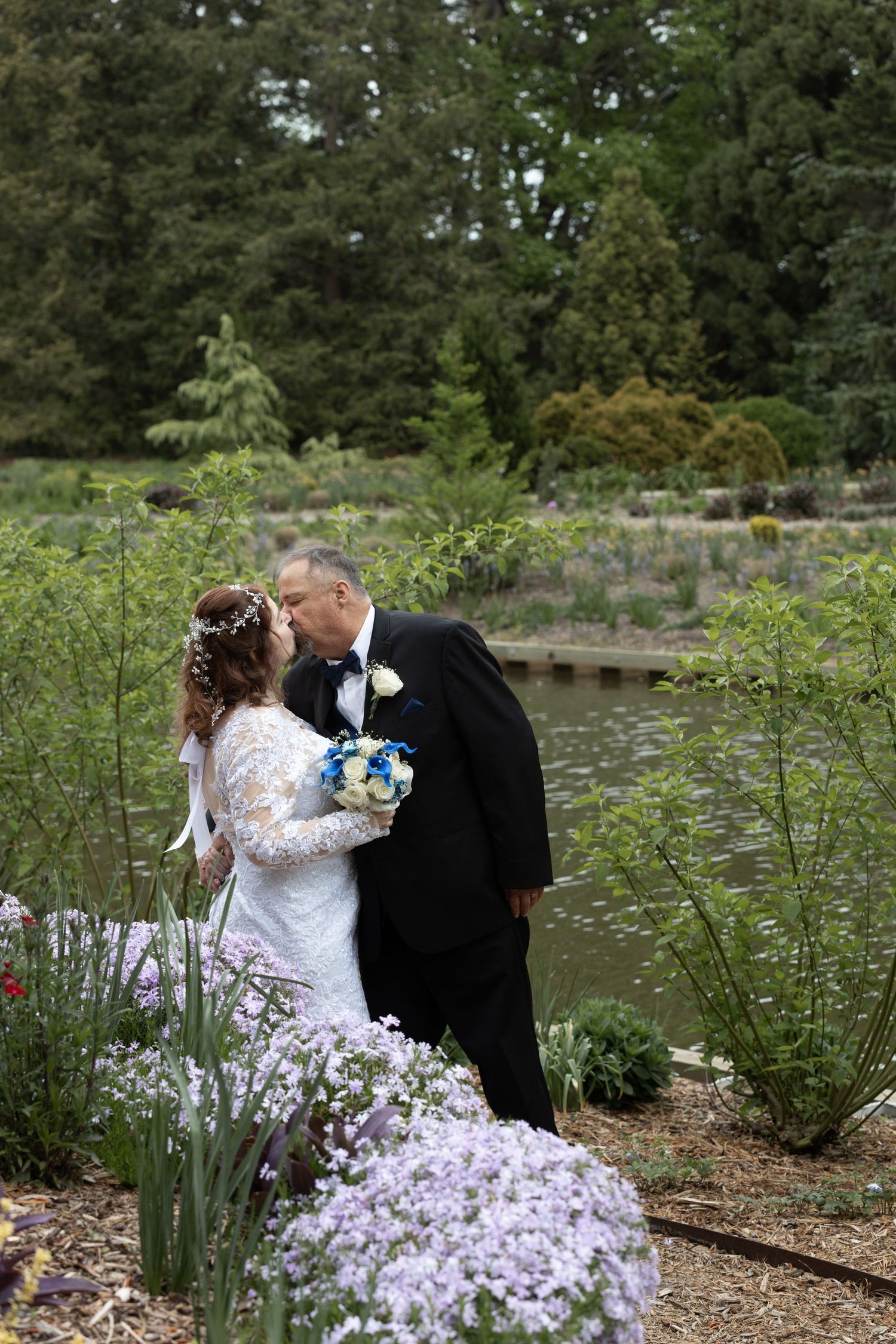 Bride and groom share a romantic kiss beside a pond in a lush garden setting.