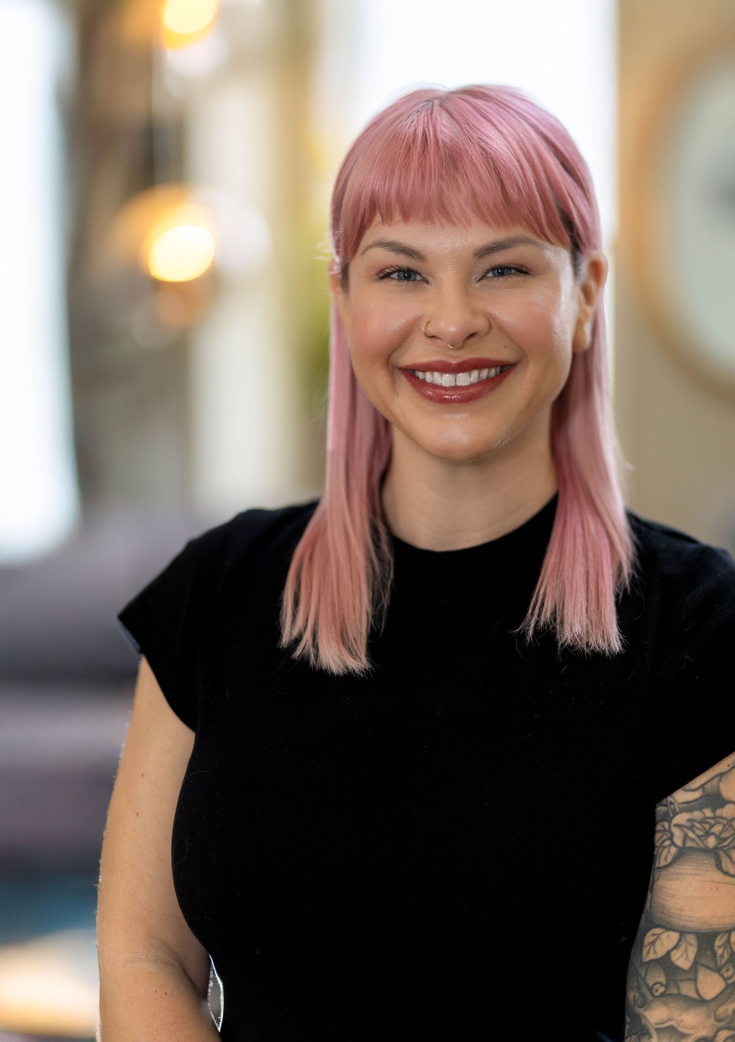 Smiling woman with pink hair and tattoos wearing black top poses for professional headshot indoors.