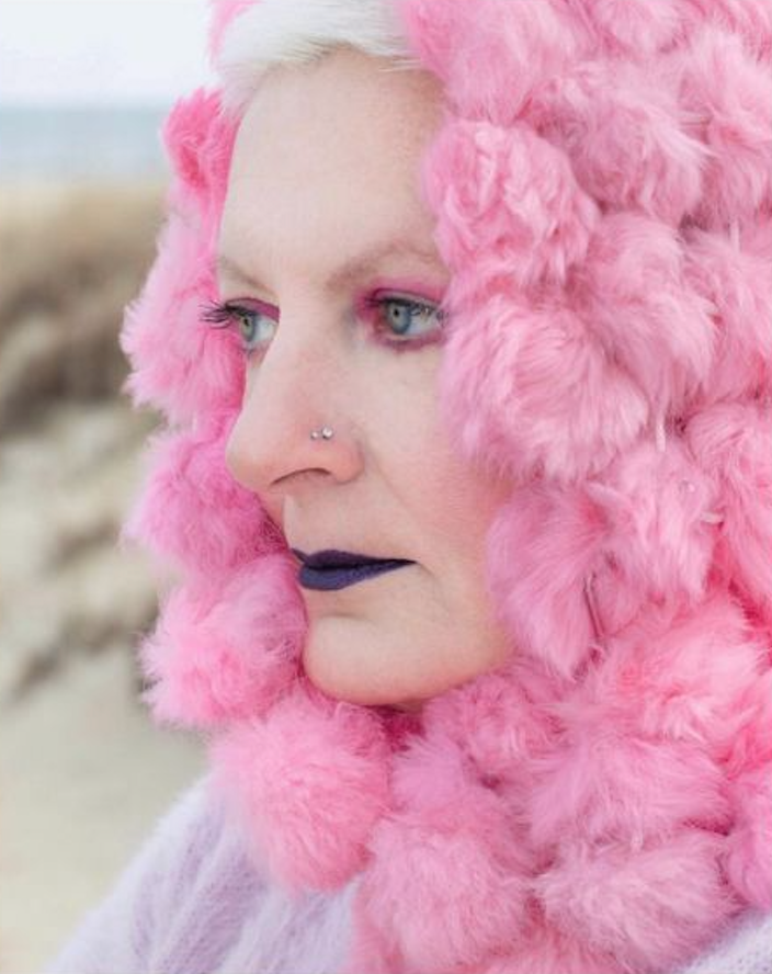 Woman wearing pink fluffy pom-pom hood with bold dark lips and green eyes at beach.