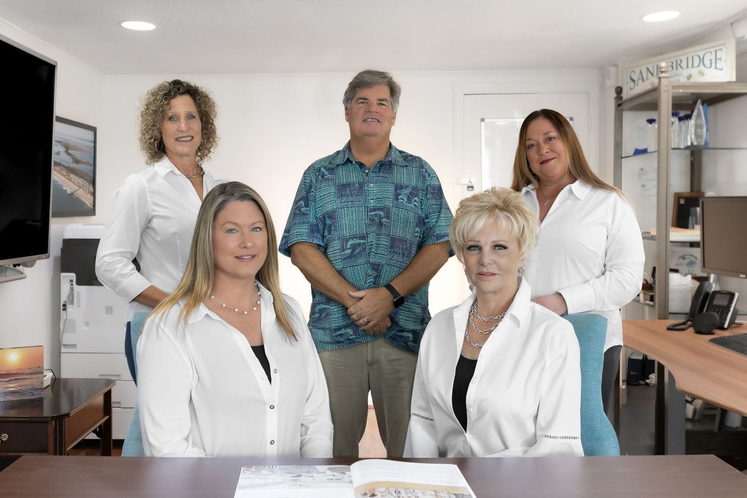 Five professional office staff members posing together in a bright workspace, wearing white and casual business attire.