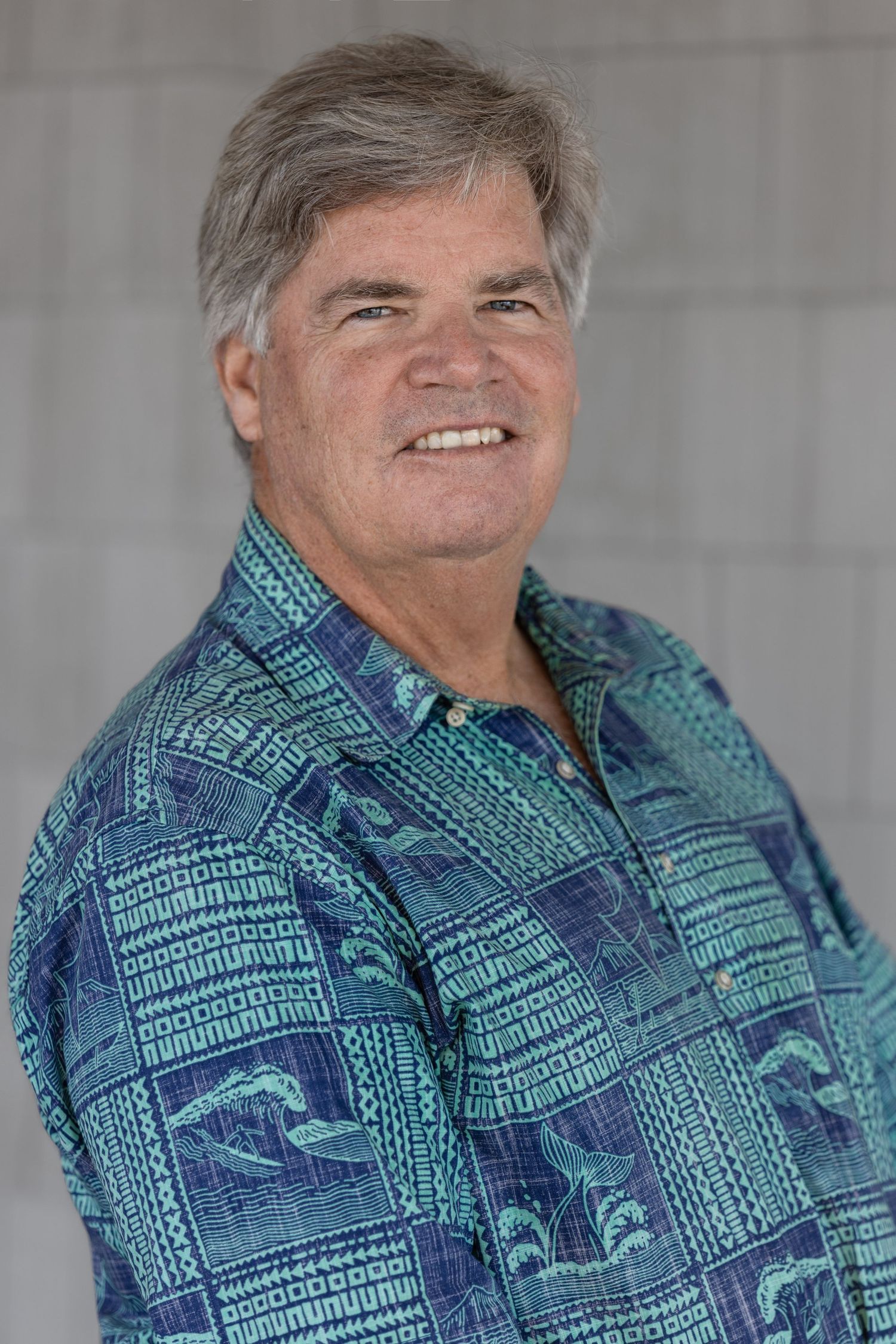 Smiling middle-aged man with gray hair wearing a blue tropical Hawaiian shirt, standing outdoors.
