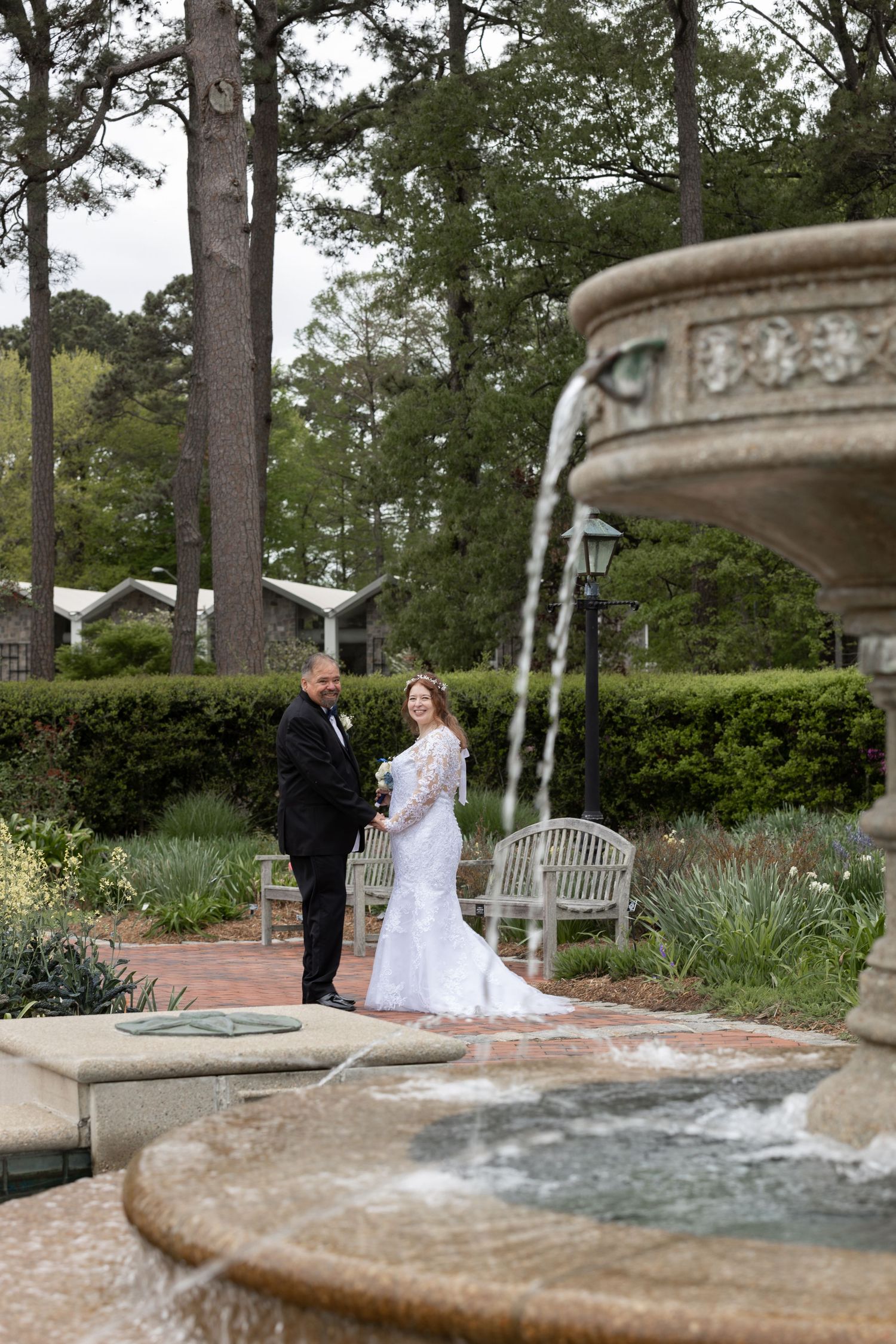 Bride and groom holding hands near decorative stone fountain in lush garden setting with tall pine trees.
