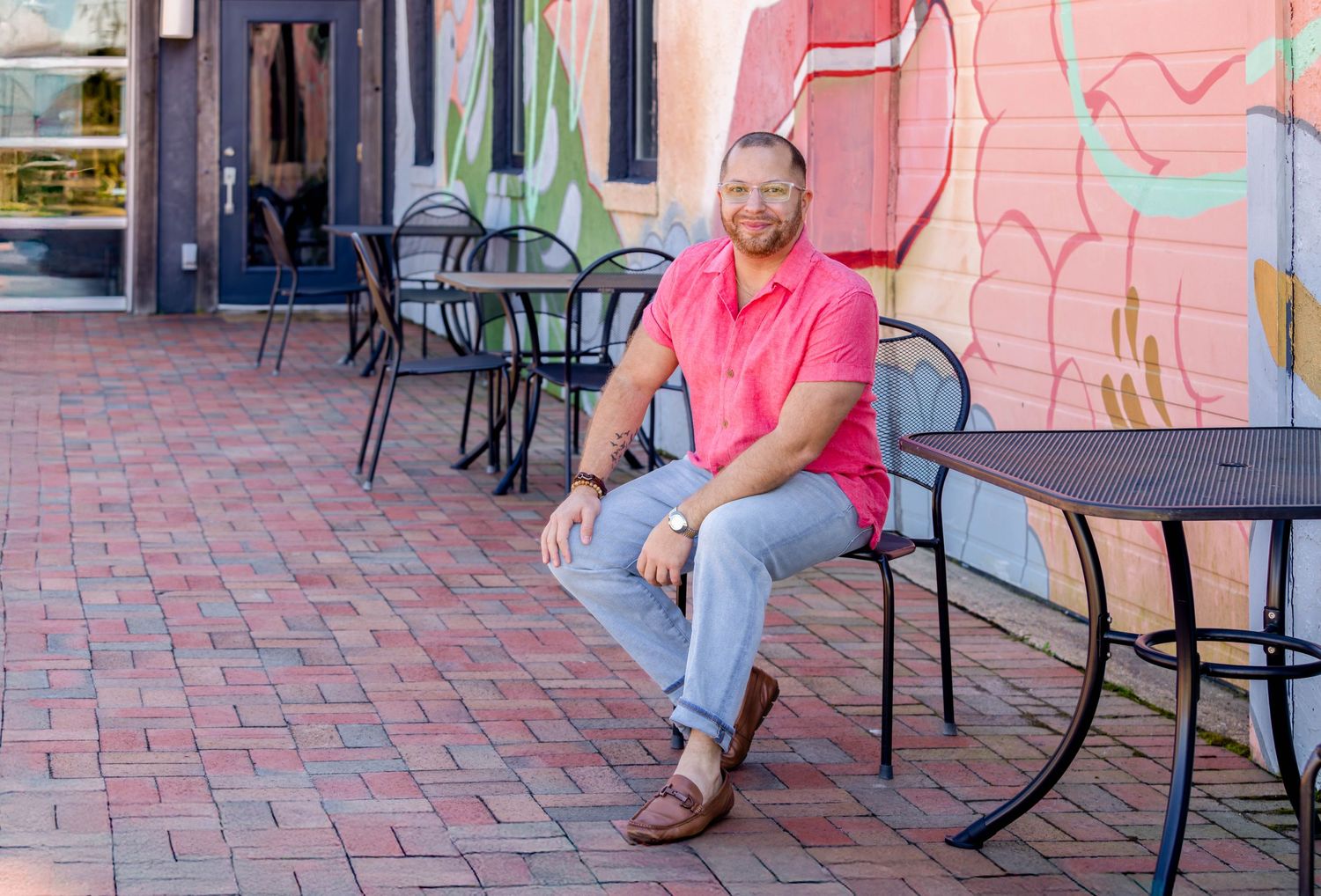 Smiling man in pink polo shirt sitting on bench outside colorful mural on brick patio.