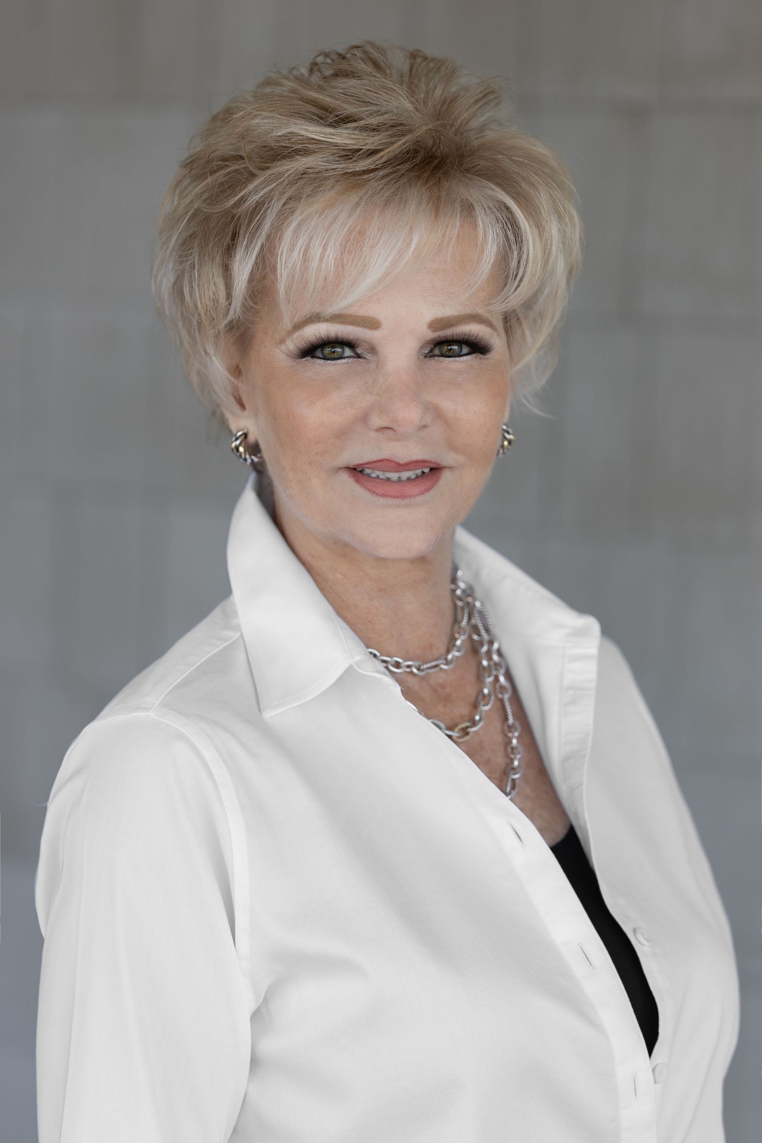 Smiling blonde woman with short hair wearing a white button-up shirt and silver necklace poses professionally.