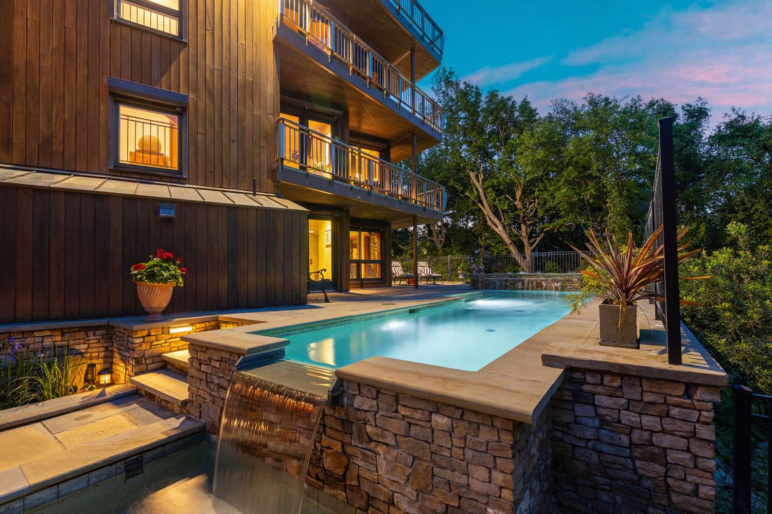 Stunning outdoor pool with stone waterfall feature beside a modern wood home at twilight.