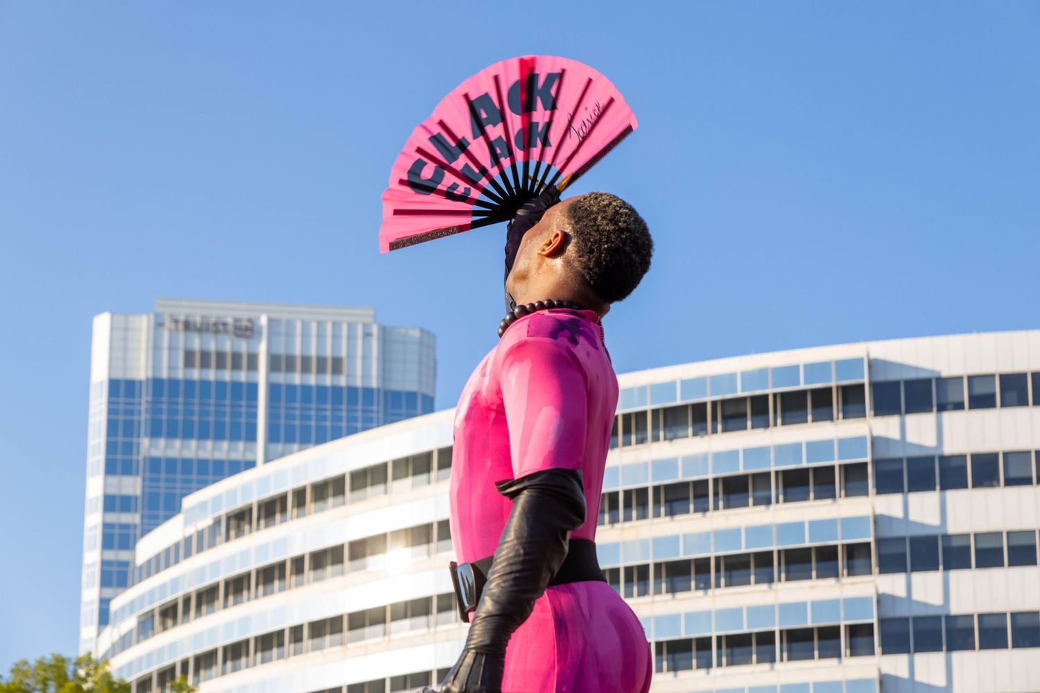 Person in pink bodysuit holding pink fan overhead against blue sky and city buildings.