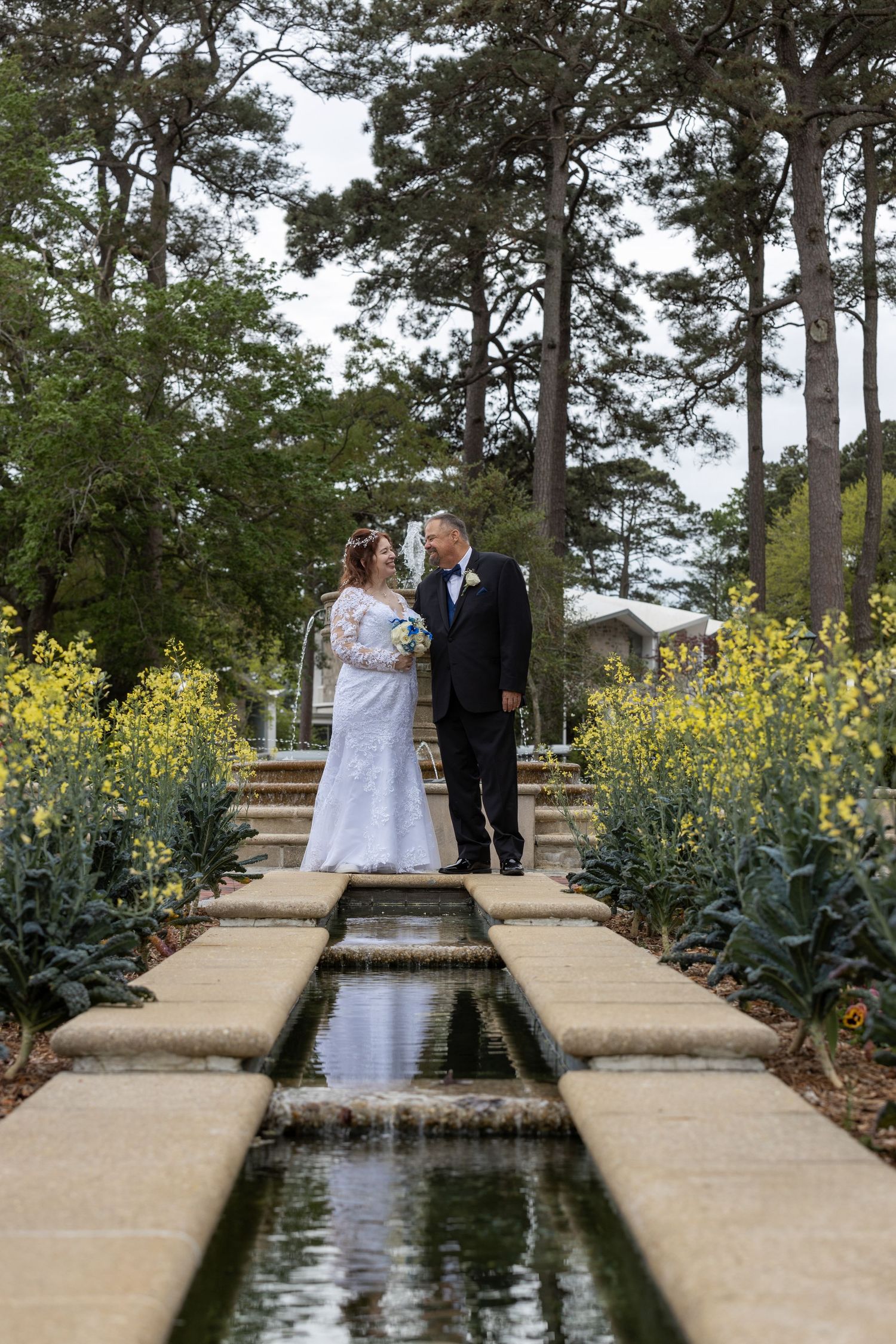 Bride and groom sharing romantic moment beside reflecting pool pathway surrounded by yellow flowers and tall pines.
