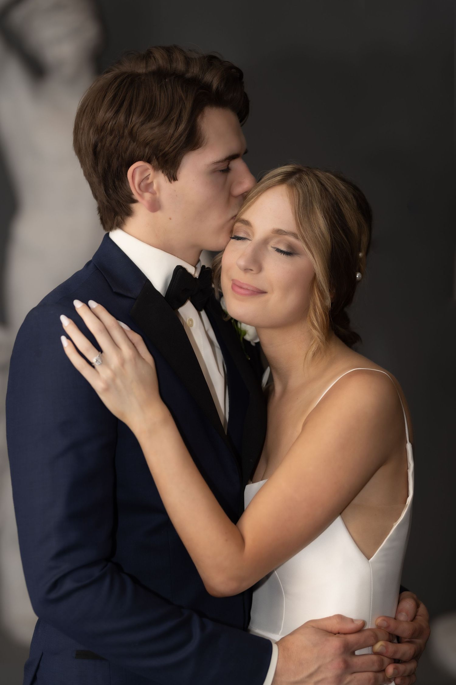 Groom in navy tuxedo tenderly kisses bride in white slip dress during romantic wedding portrait session.