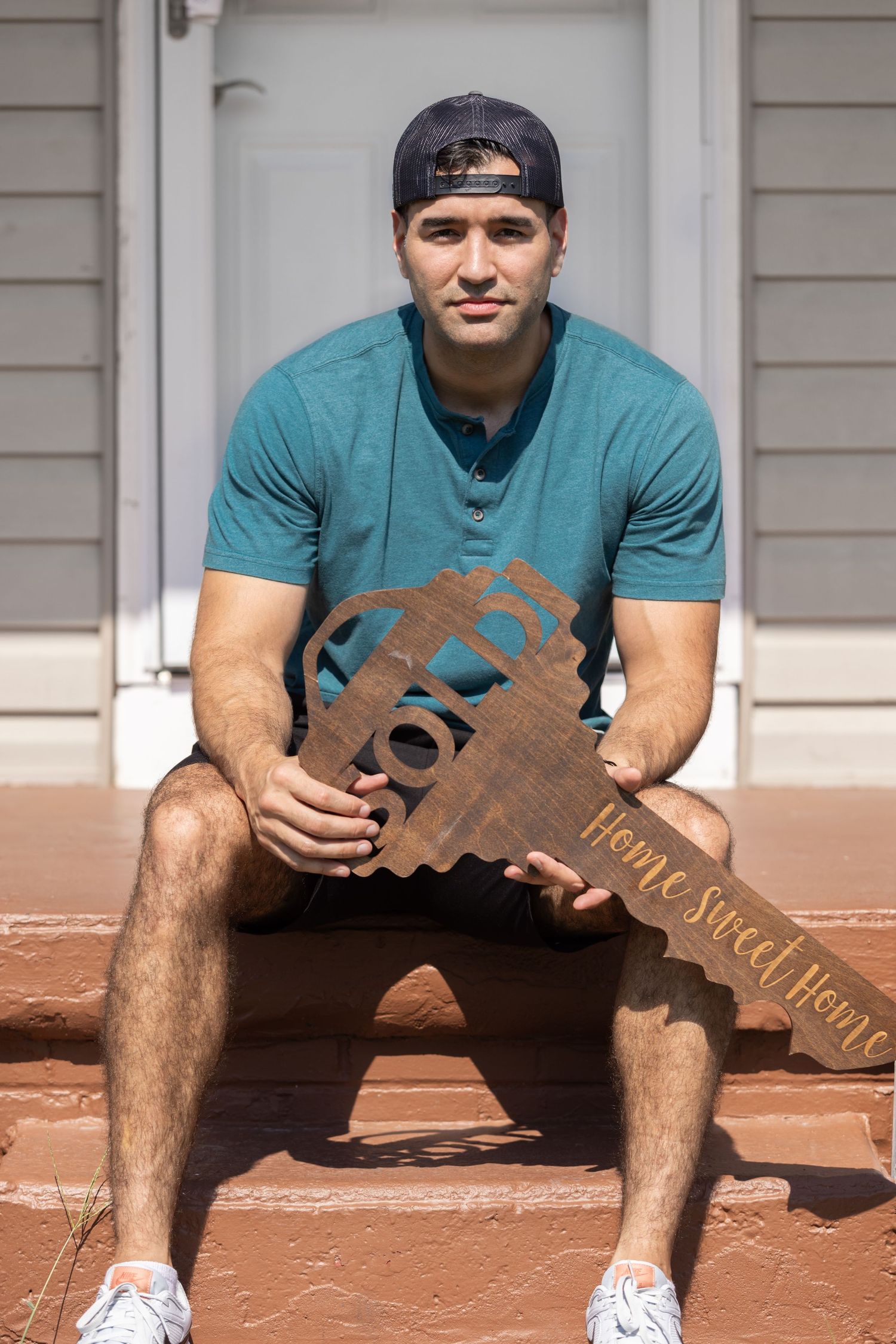 Young man sitting on porch steps holding a wooden Home Sweet Home key sign.
