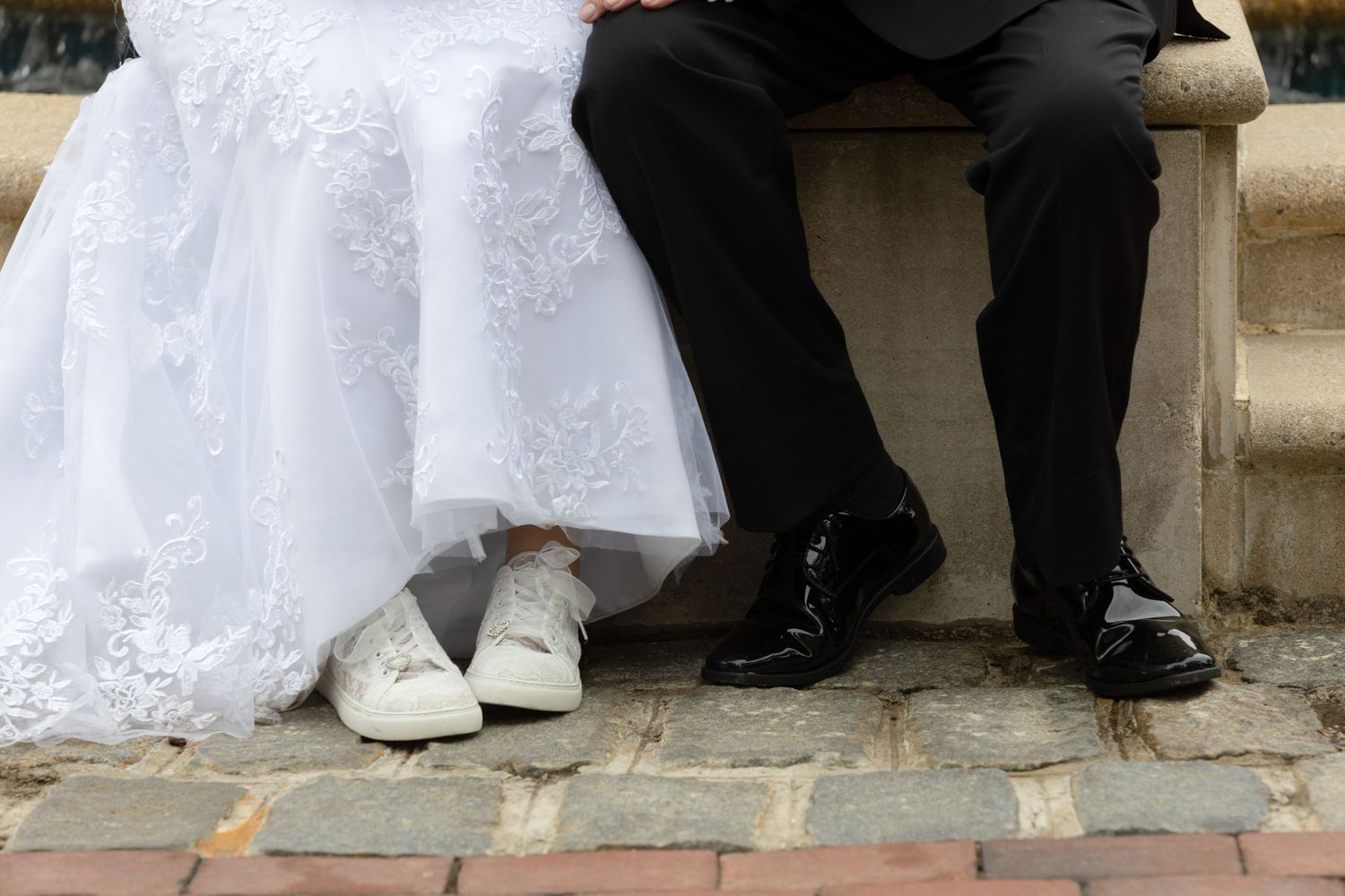 Close-up of bride in white lace dress and sneakers sitting beside groom in black suit on wedding day.