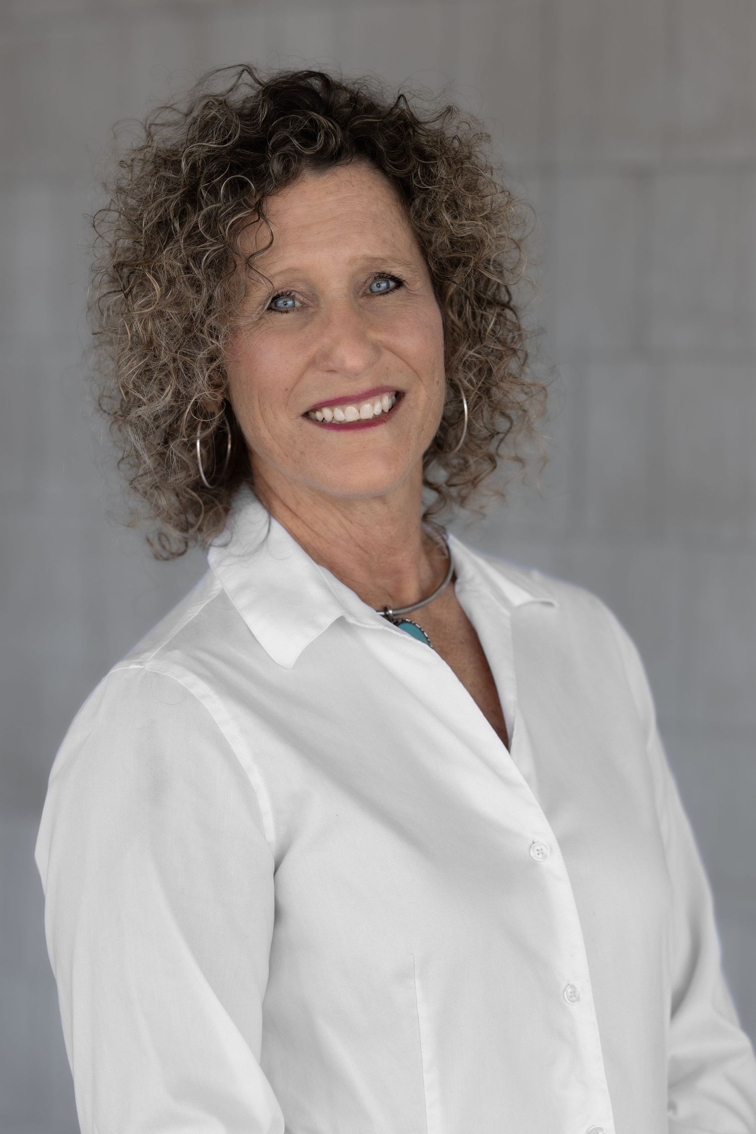 Smiling middle-aged woman with curly hair wearing a white button-up shirt and turquoise necklace against a gray background.