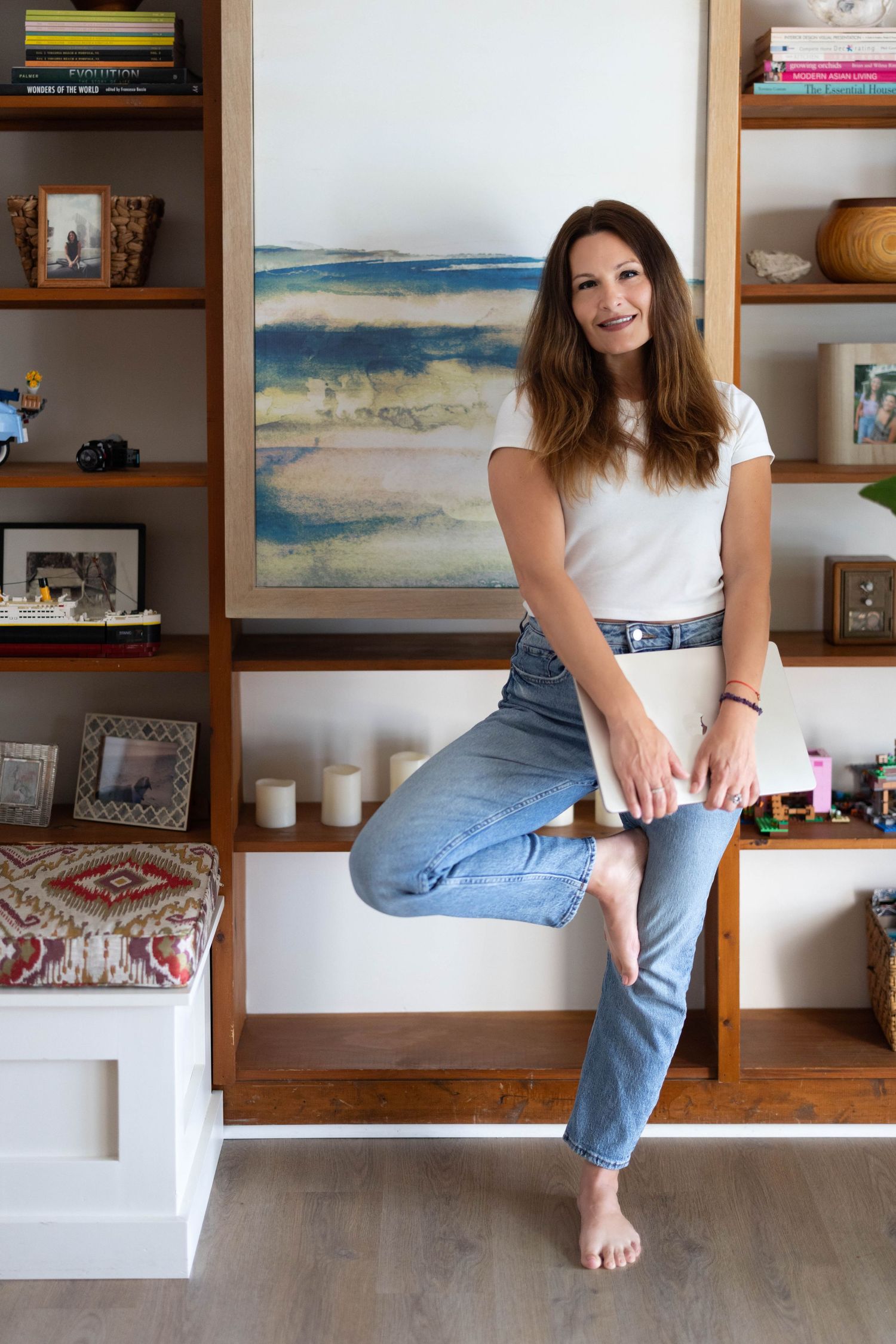 Brunette woman in white tee and jeans sitting on built-in shelf with coastal artwork and decor in background.