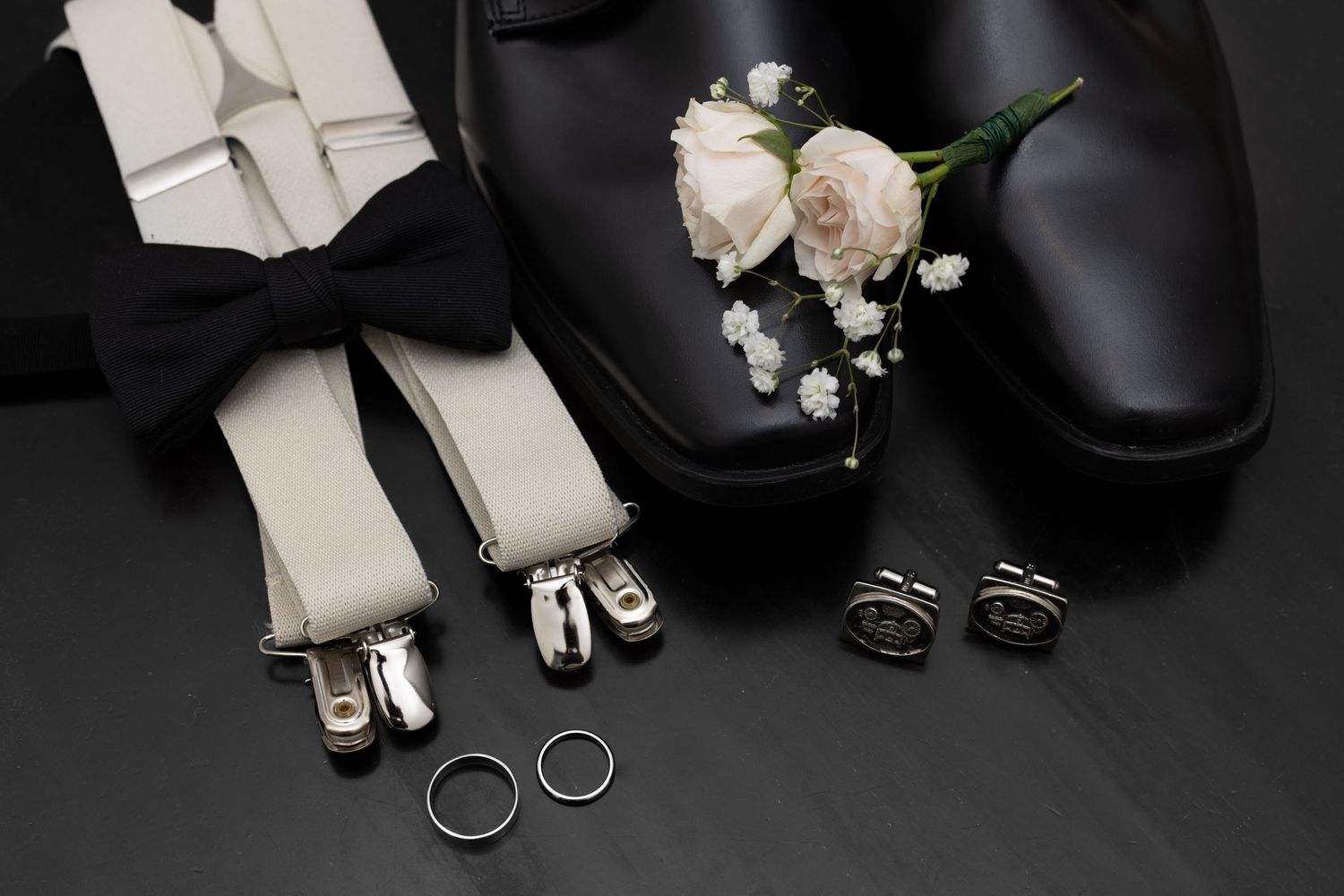 Elegant groom wedding accessories including bow tie, suspenders, rings, cufflinks, and boutonniere on dark surface.