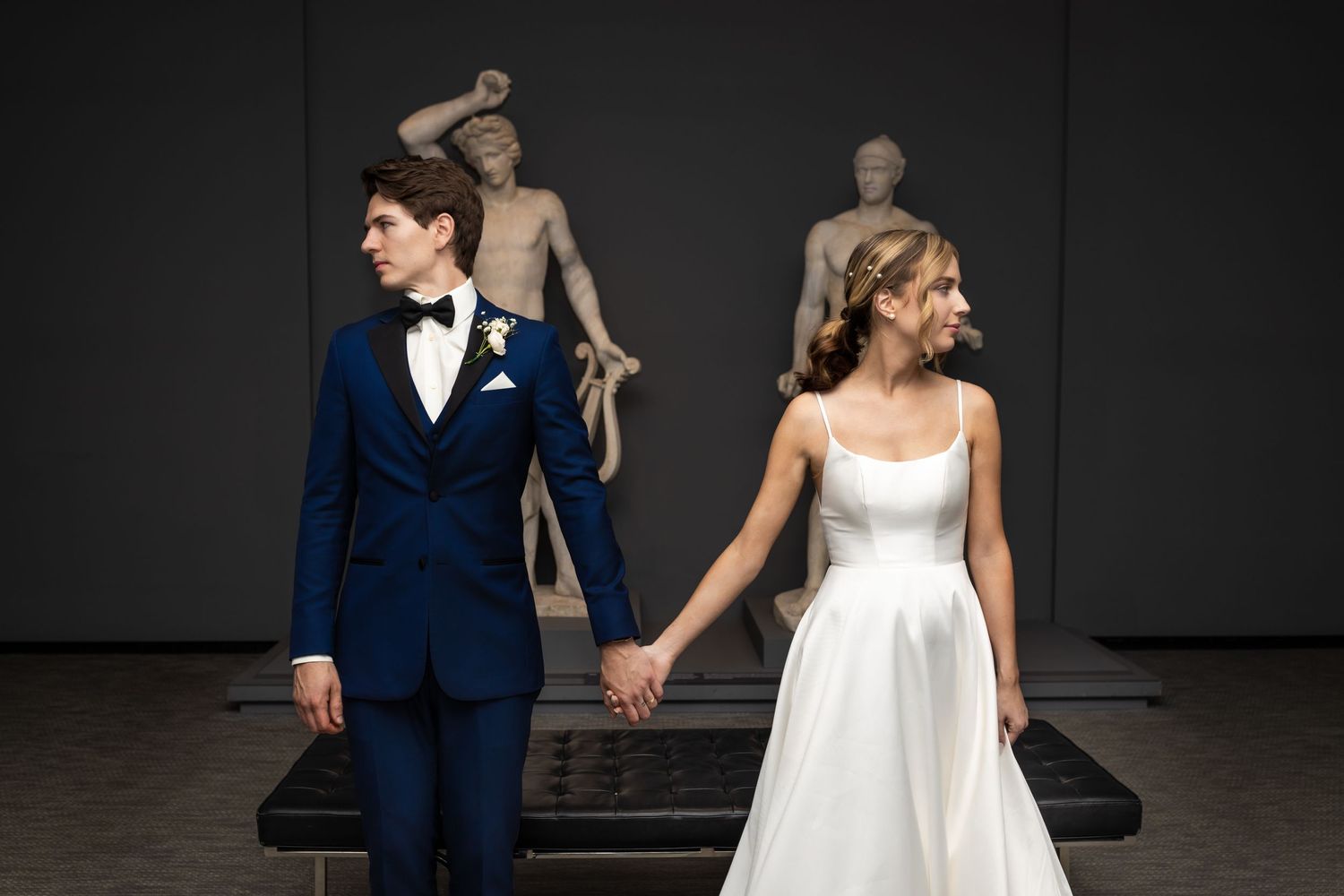Bride in white gown and partner in navy suit hold hands before classical sculptures in elegant museum setting.