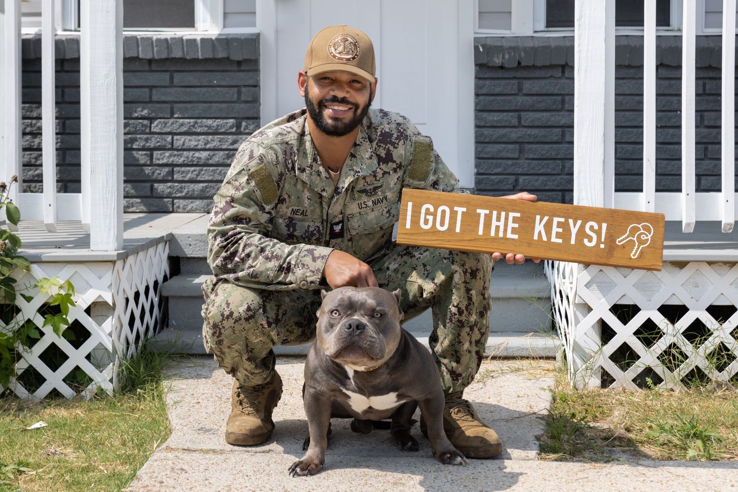 Navy serviceman in uniform crouches beside gray bulldog holding an I Got The Keys sign outside new home.