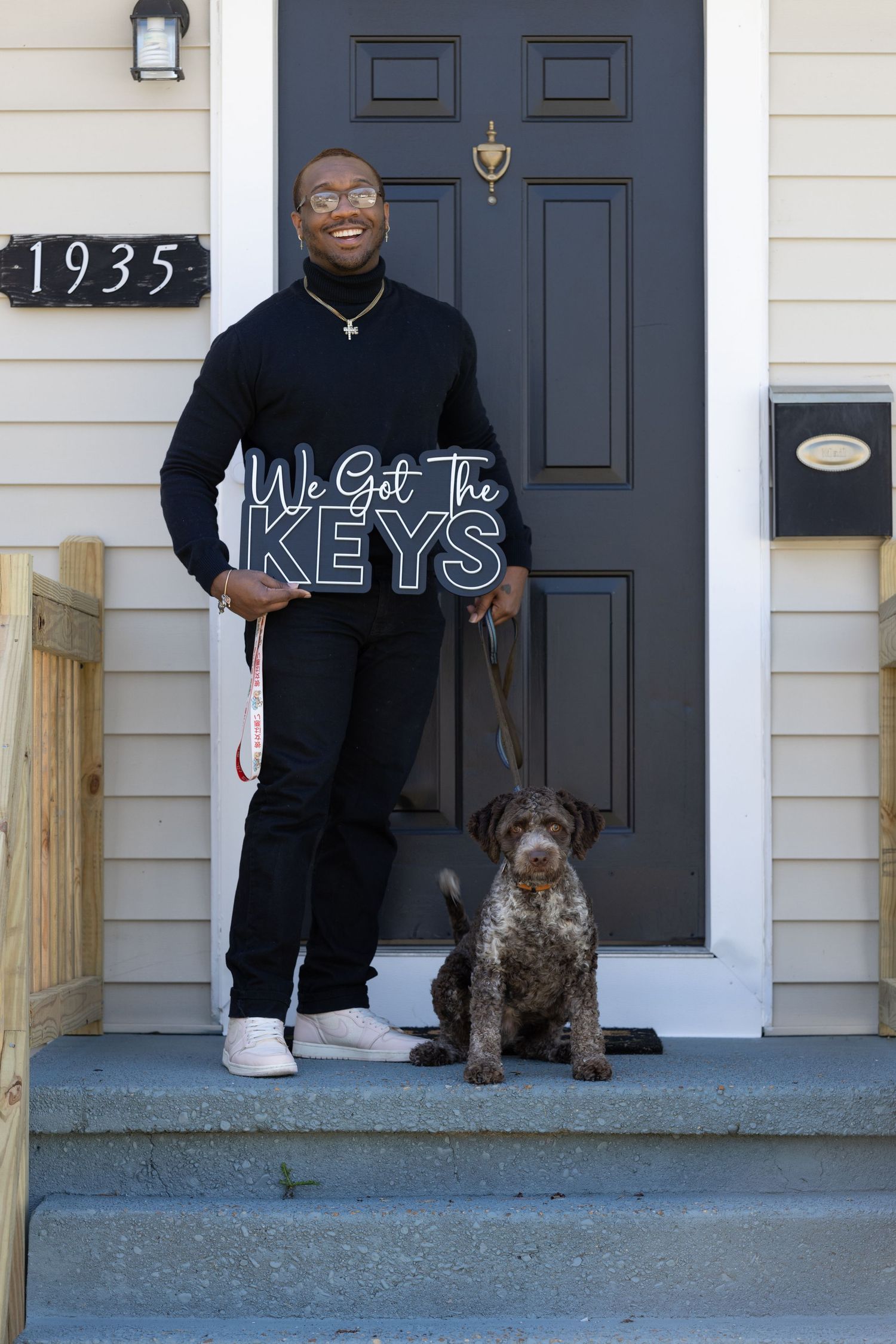 Man holding 'We Got The Keys' sign with dog on steps of new home numbered 1935, celebrating homeownership.