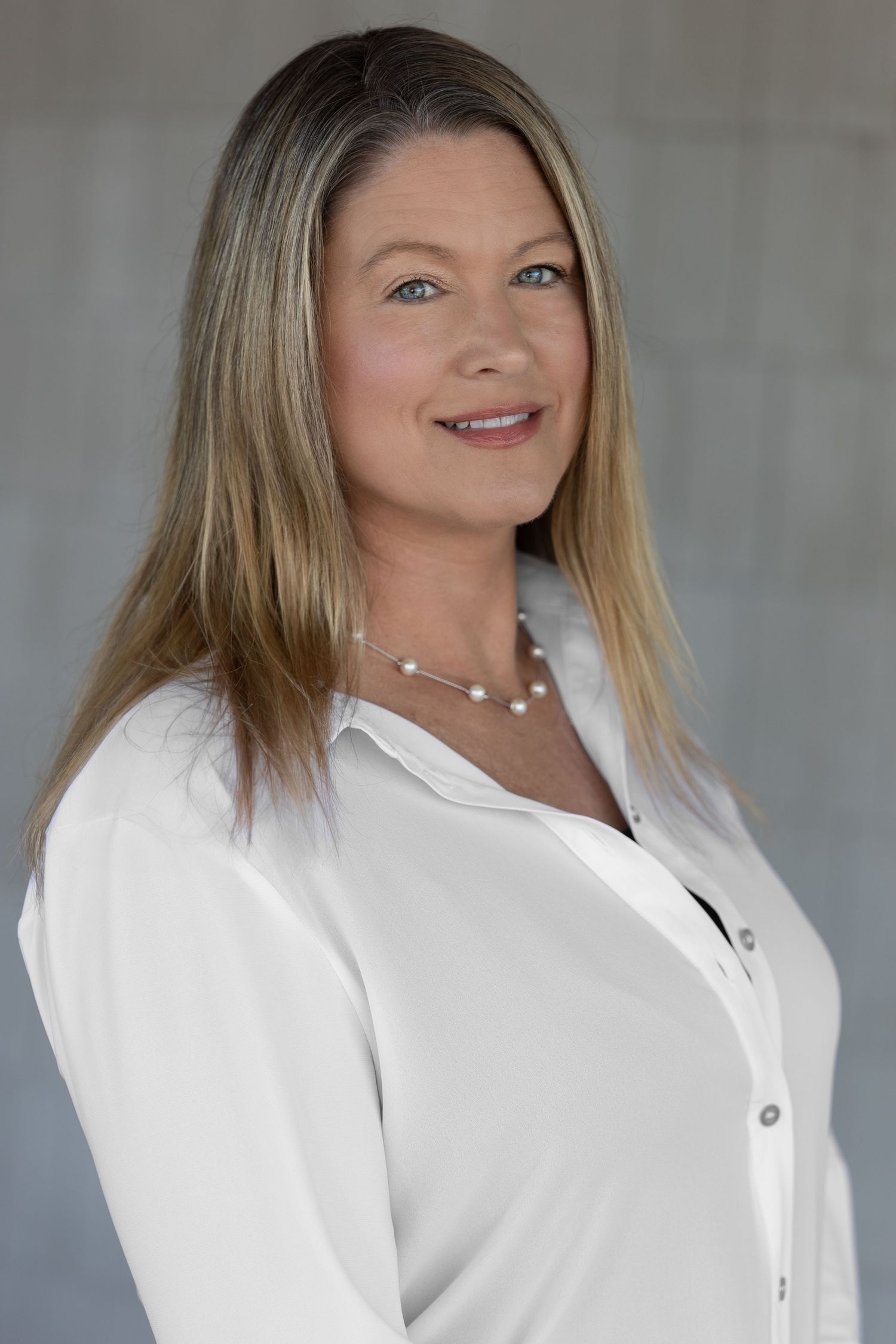 Professional woman with blonde hair wearing a white blouse and pearl necklace, smiling confidently in a studio setting.