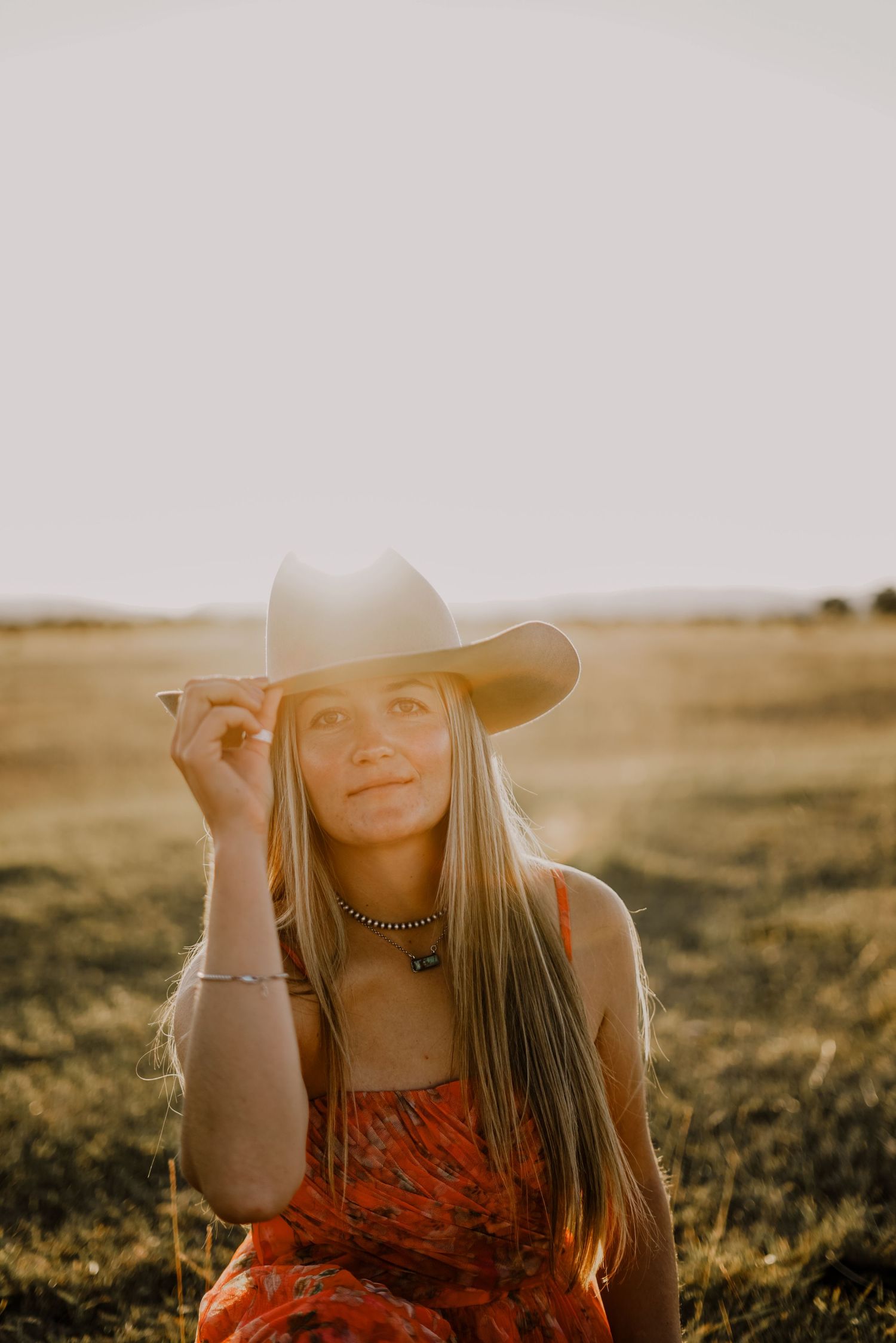 A young woman in a pink dress wearing a cowboy hat  holding the brim with her hand. A sunset is setting behind mountains in the background