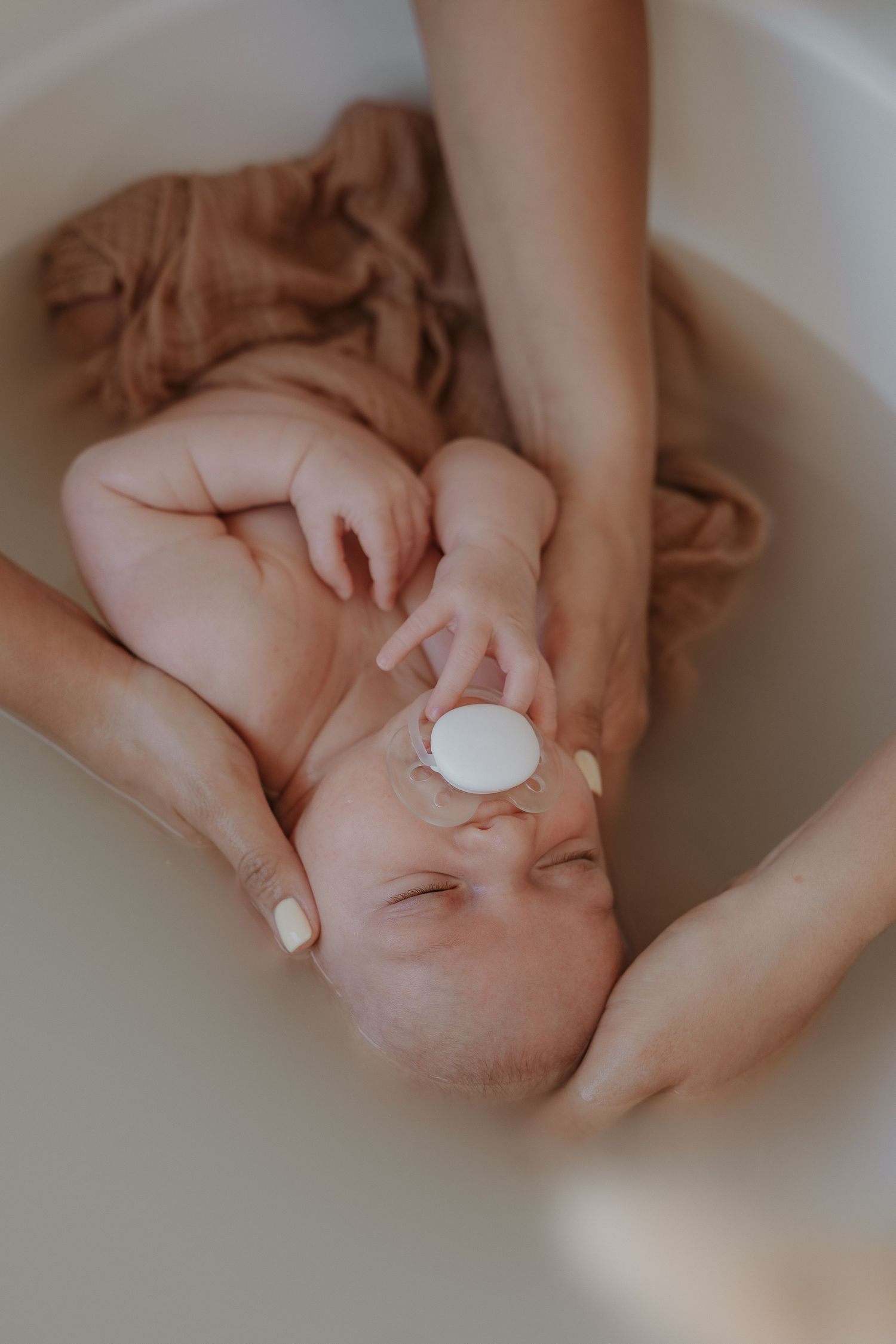 Newborn baby being gently bathed in a white tub with a pacifier, supported by caring adult hands.