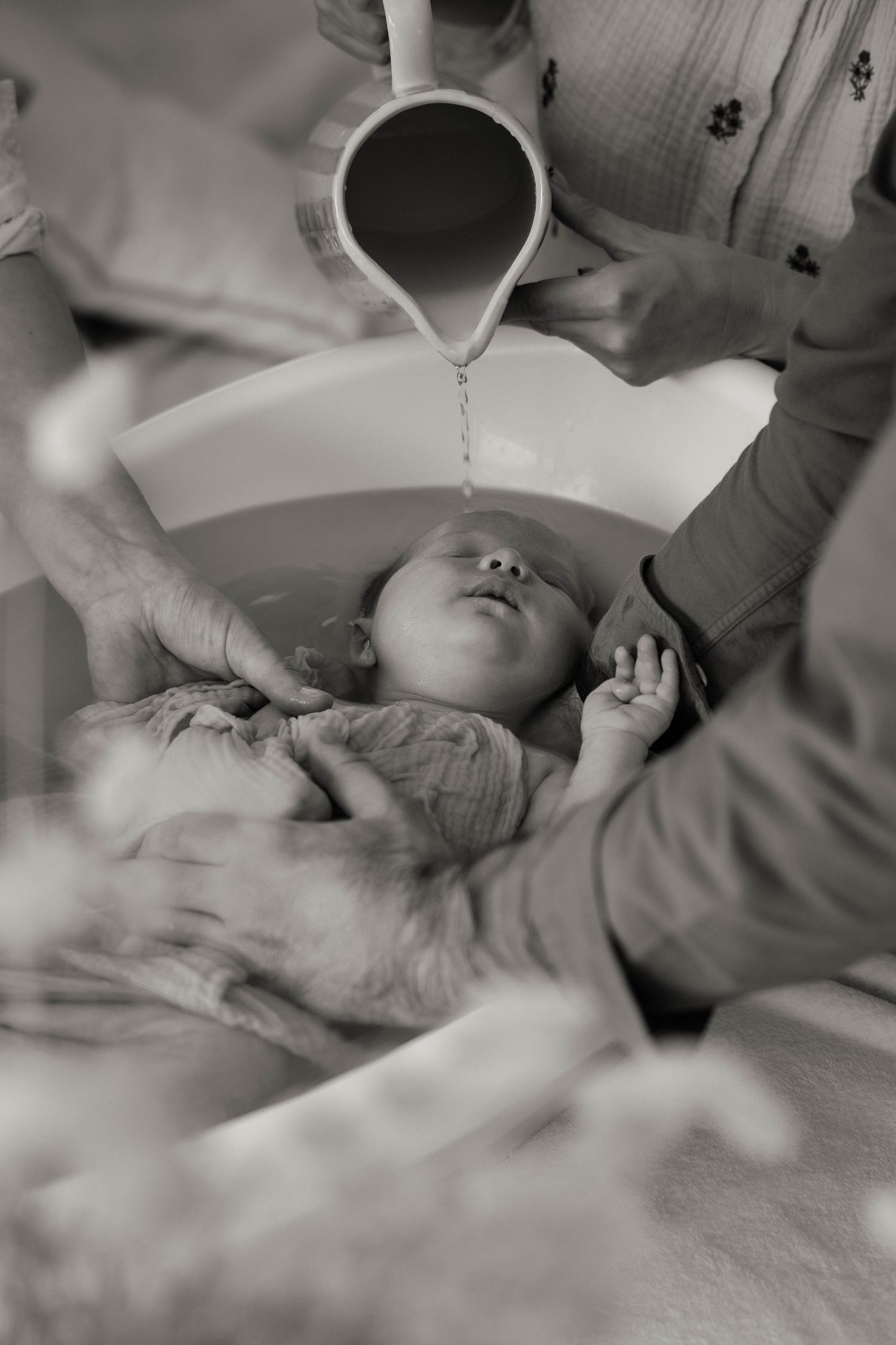 Black and white photo of a newborn baby being baptized with water poured from a pitcher over their head.